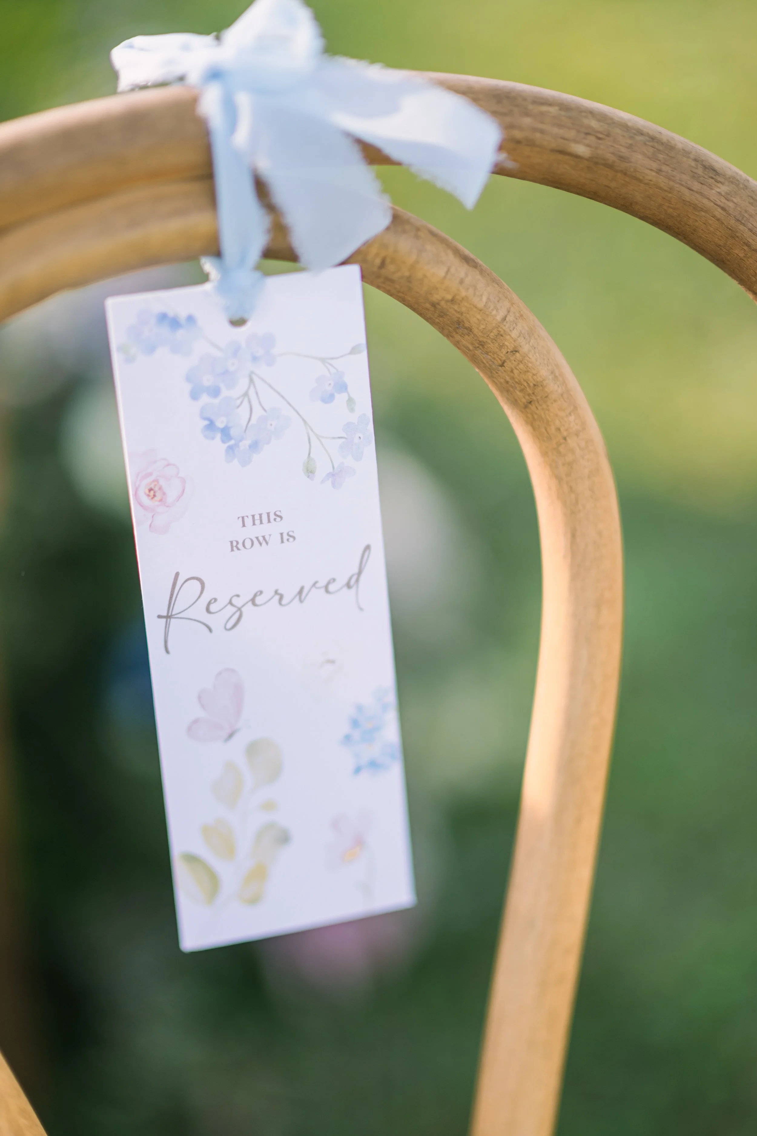 A white paper with blue and pink flower illustrations hangs from a light brown wooden chair, tied with a light blue ribbon. The paper reads 'This row is reserved' in elegant script. The background is blurred green foliage.