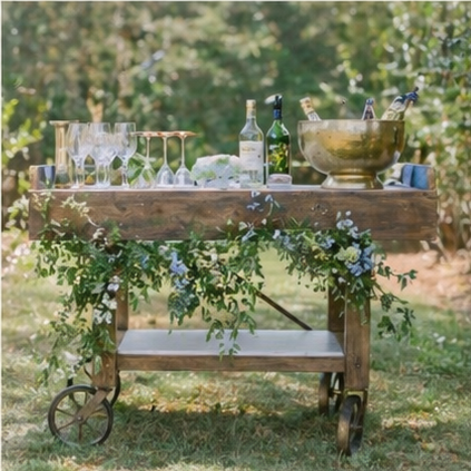 Outdoor wooden cart with glasses, bottles, and a brass bowl, decorated with greenery and flowers.