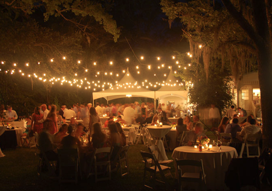 Outdoor nighttime gathering with string lights hanging above tables filled with people, lit by candles, under trees and tents.