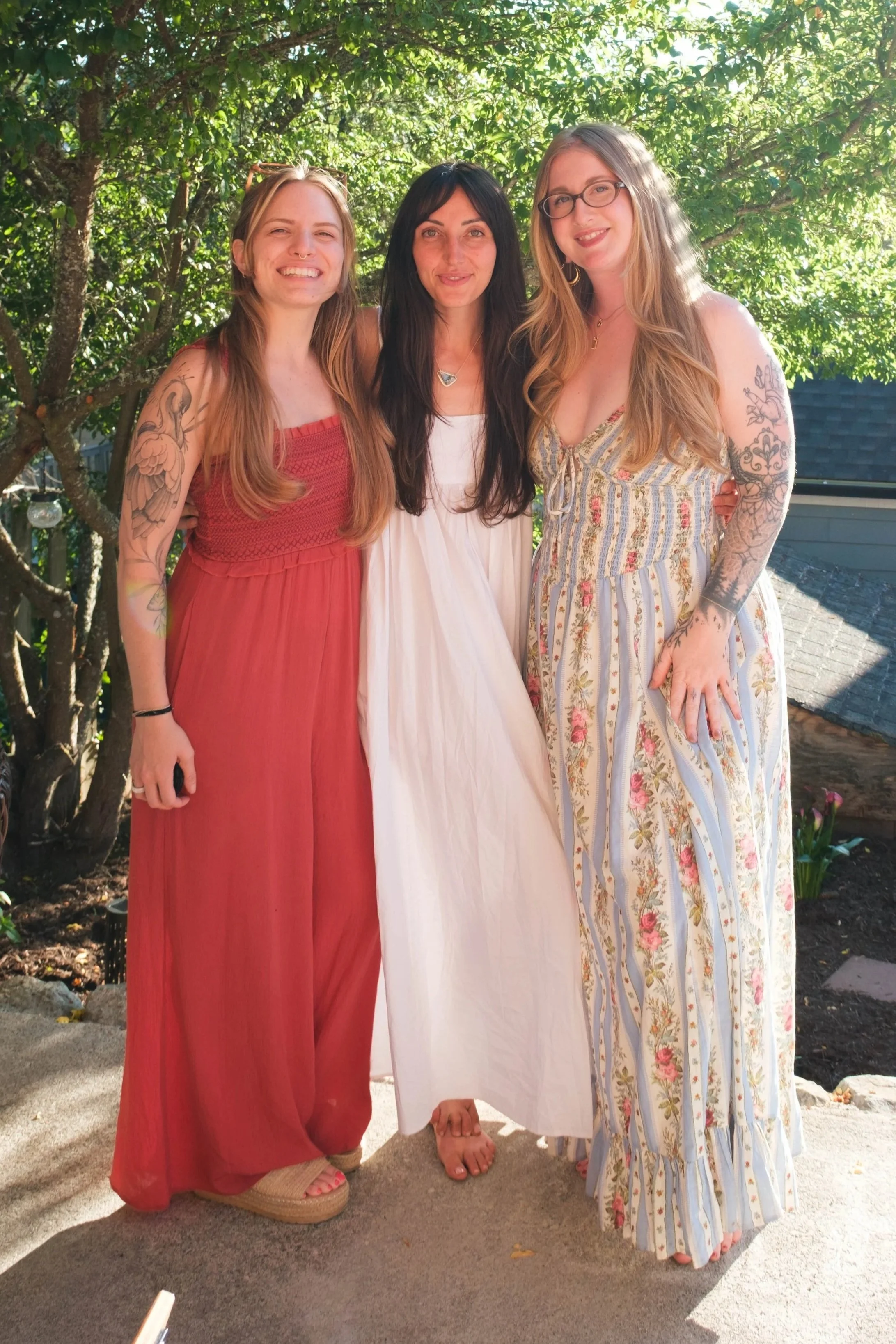 Three women standing together outdoors, smiling, with lush green trees in the background. Two women have long, wavy hair and tattoos on their arms, and the woman in the middle has straight dark hair and is wearing a white dress. The woman on the left wears a red dress, the woman on the right wears a light-colored, patterned maxi dress, and the woman in the middle wears a white dress. All are barefoot.