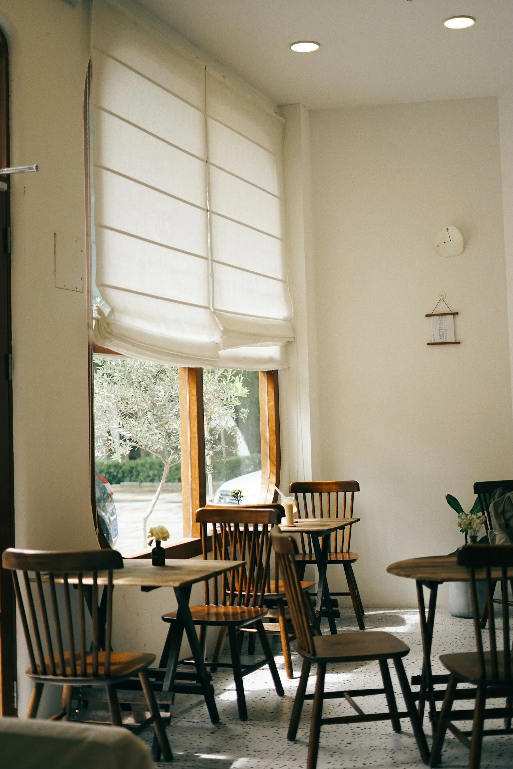 Interior of a cozy cafe with wooden chairs and tables, sunlight coming through a large window with white Roman blinds, and a small flower on each table.