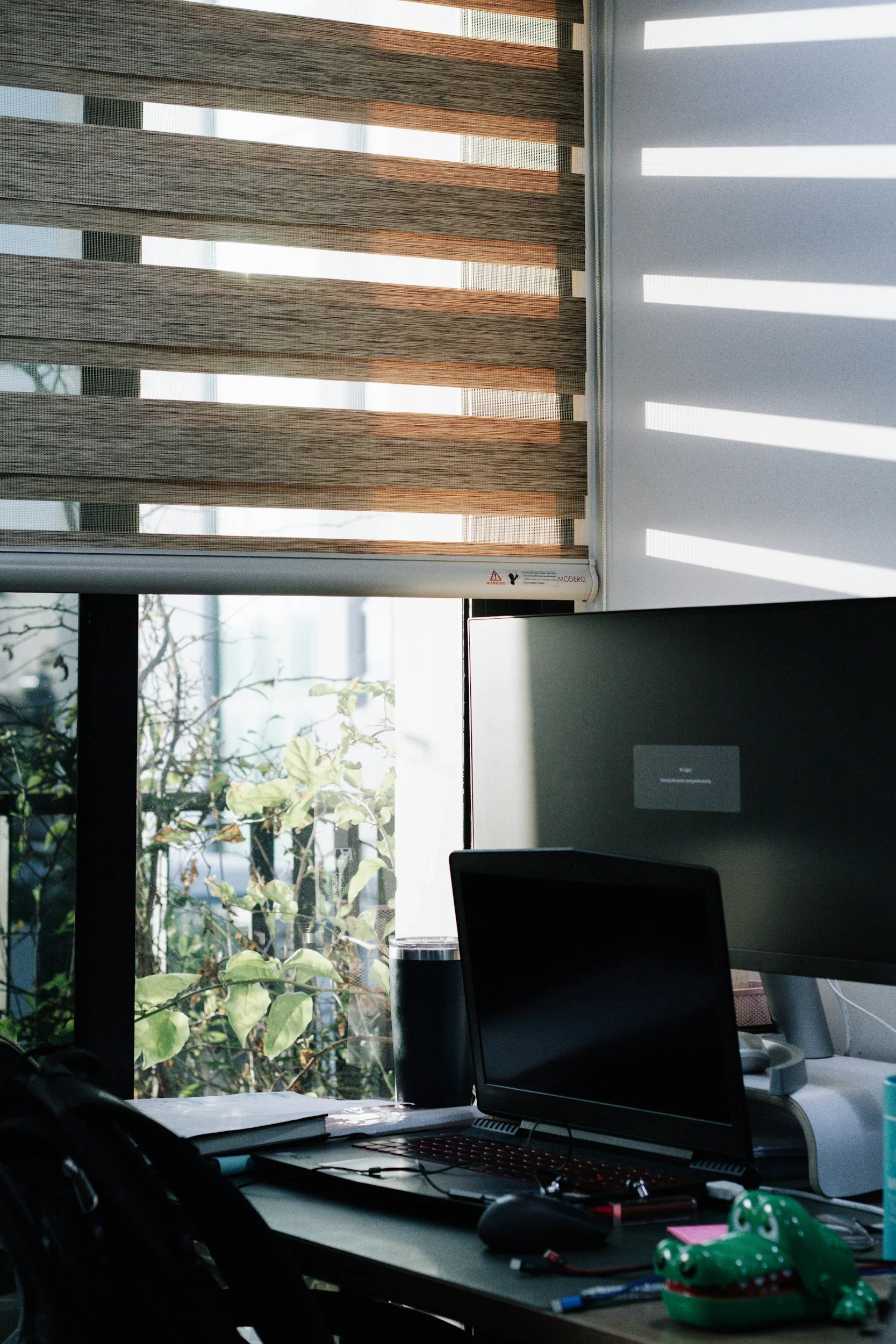 A cluttered office desk with a laptop, mouse, notebooks, pens, and a green crocodile-shaped toy in front of a window with brown horizontal blinds and sunlight streaming through.