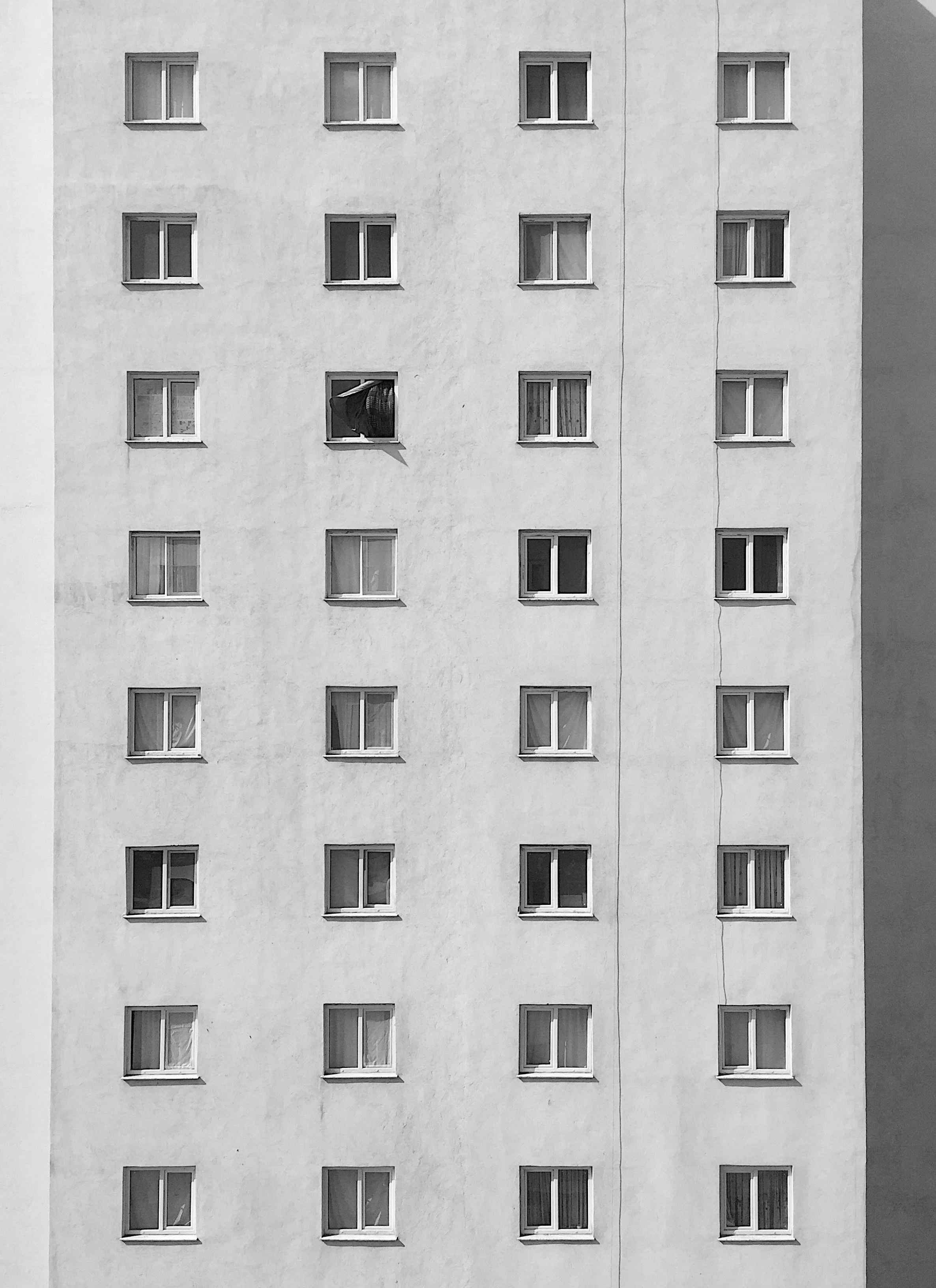 A black and white photo of a multi-story apartment building with 20 rectangular windows arranged in a grid. One window has a partially open curtain and hanging laundry outside.