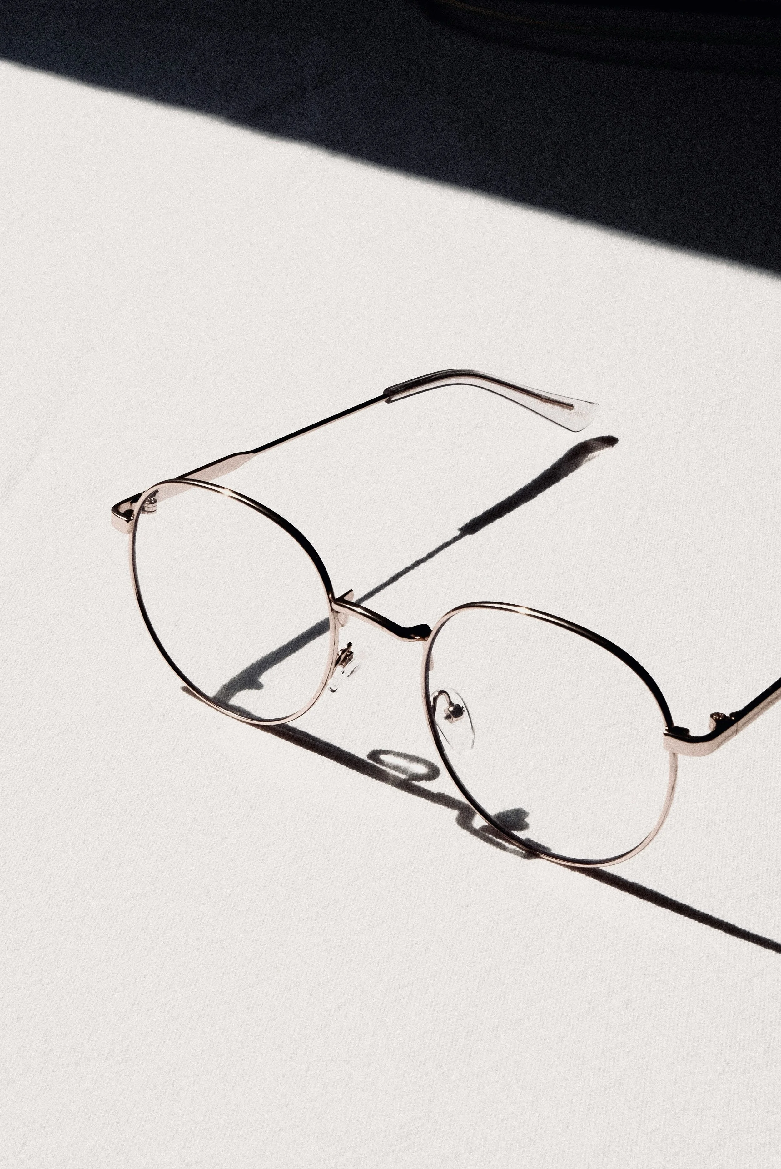 A pair of round eyeglasses with thin metal frames rests on a white surface with a shadow cast nearby.