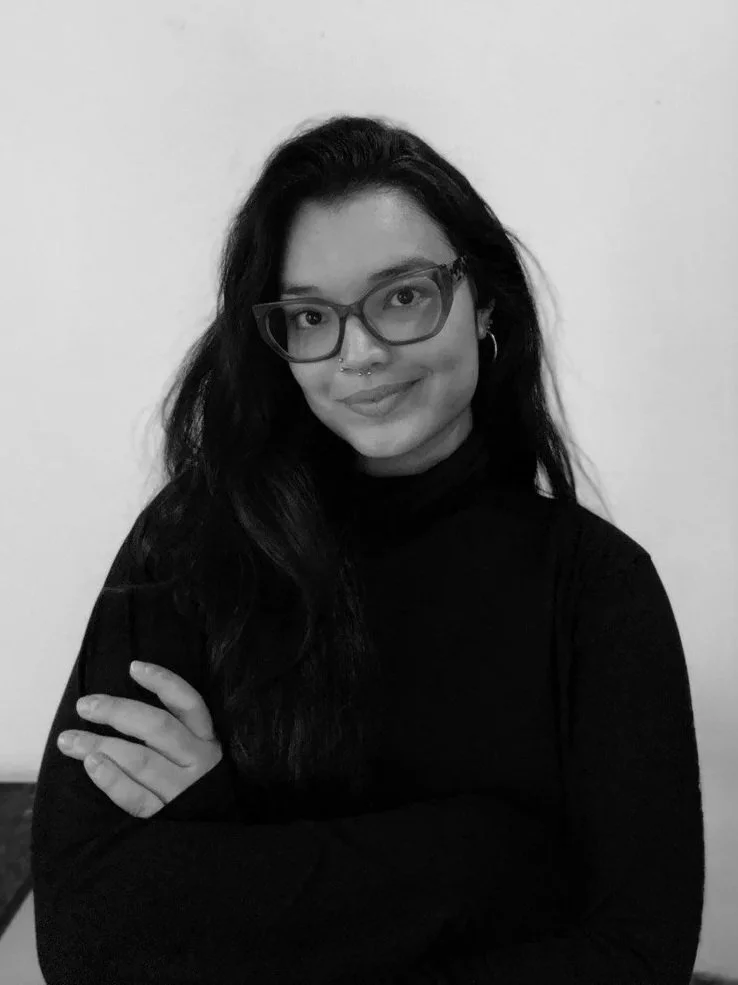 A woman with long dark hair, glasses, nose ring, wearing earrings and a black turtleneck, smiling with arms crossed against a plain white wall.