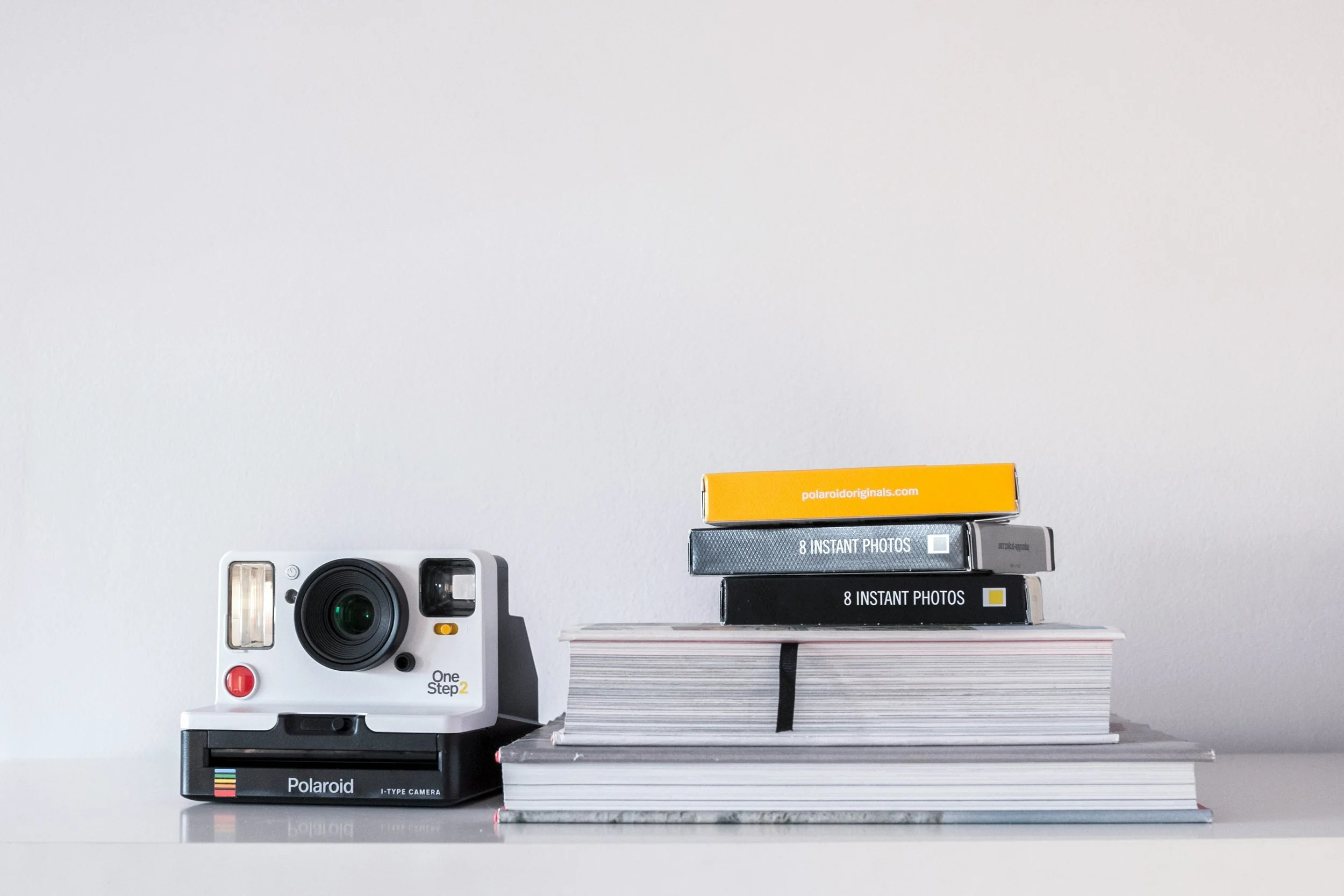 A stack of photo film boxes, a Polaroid camera, and a vintage instant camera, all arranged on a white surface against a plain white wall.