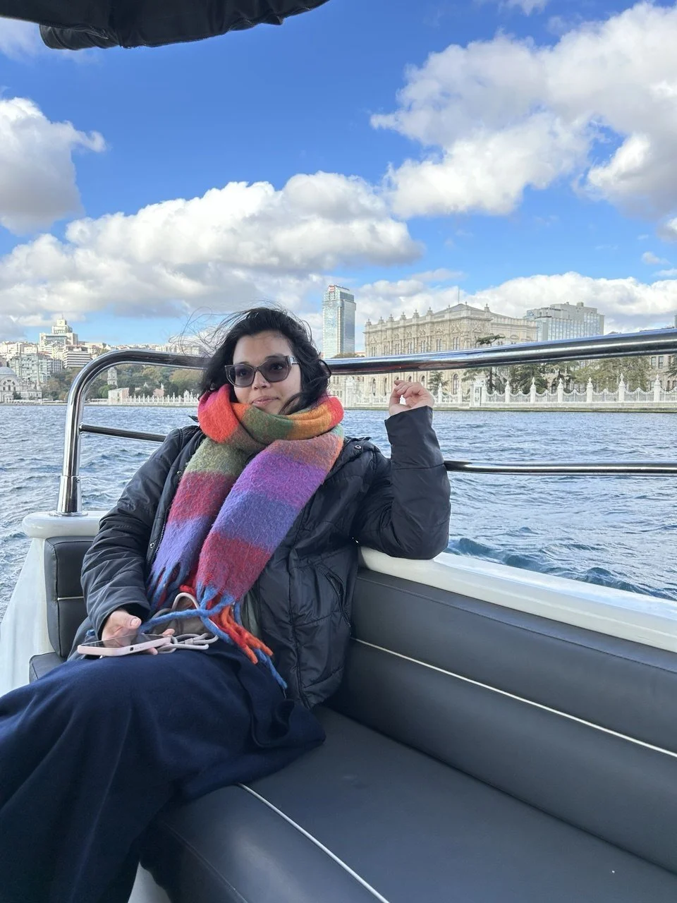 Woman sitting on a boat, wearing sunglasses, a black jacket, and a colorful scarf, with city buildings and a partly cloudy sky in the background.