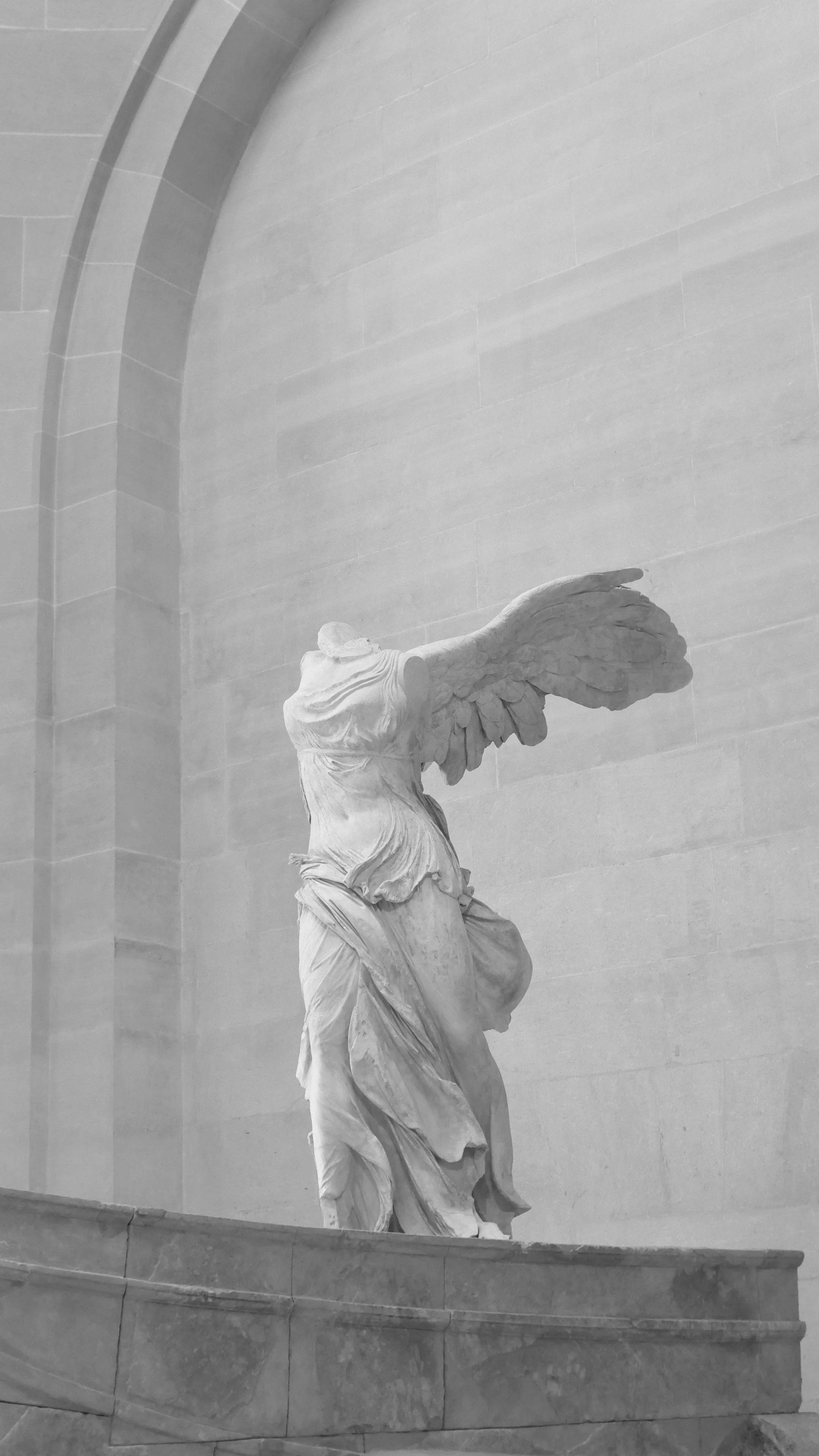 A marble sculpture of a winged woman with a flowing dress, missing her head, displayed in a grand hall with light colored stone walls and archways.
