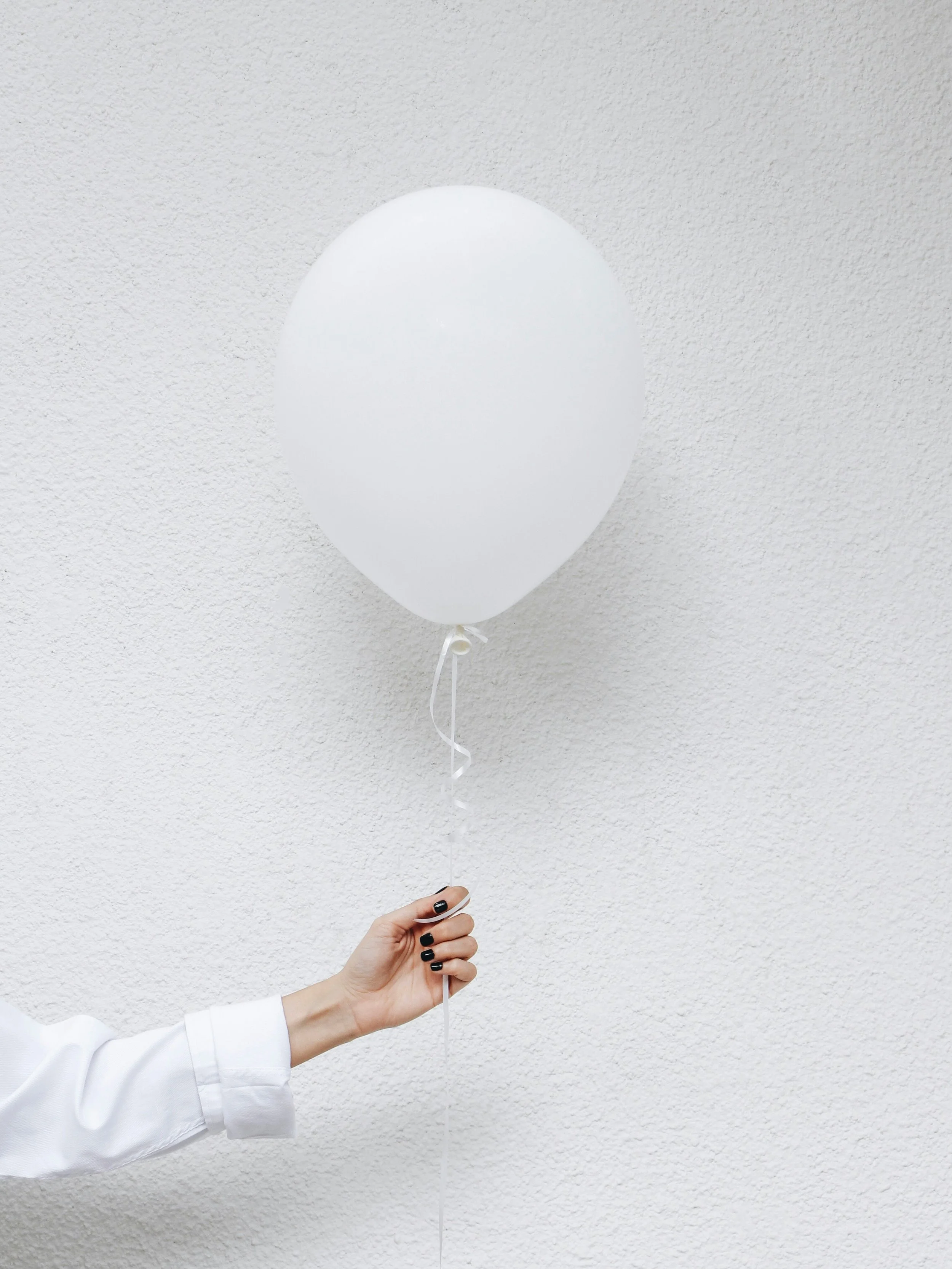 A person holding a white balloon tied to a string against a light-colored textured wall.