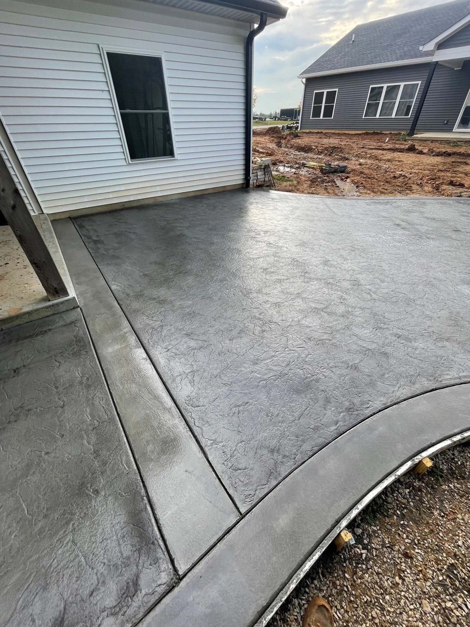View of a newly poured concrete patio next to a white house with vinyl siding, with neighboring houses and a construction site in the background.