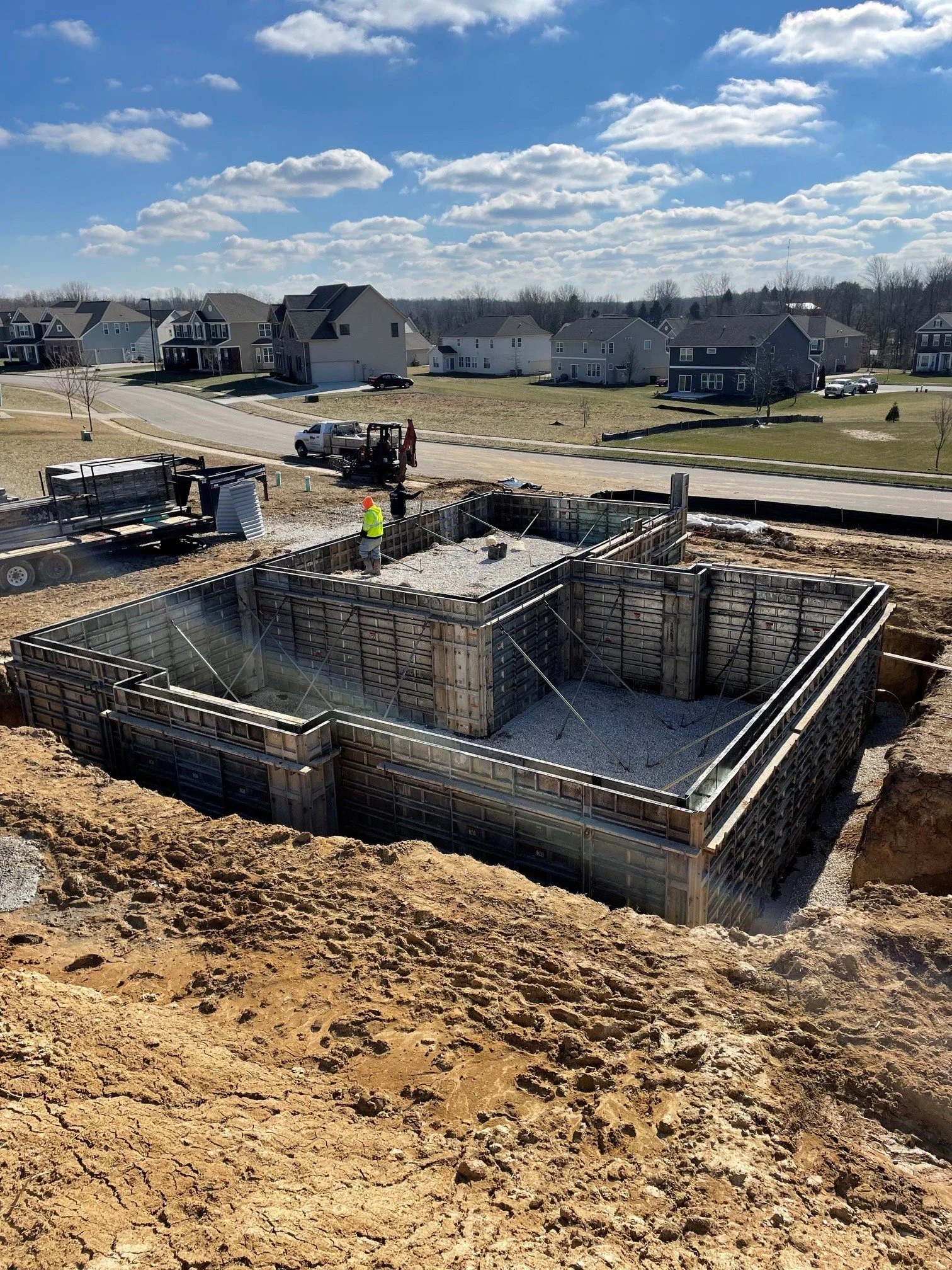 Construction site with concrete foundation and worker in safety vest and helmet, residential neighborhood with houses in the background, under a blue sky with clouds.