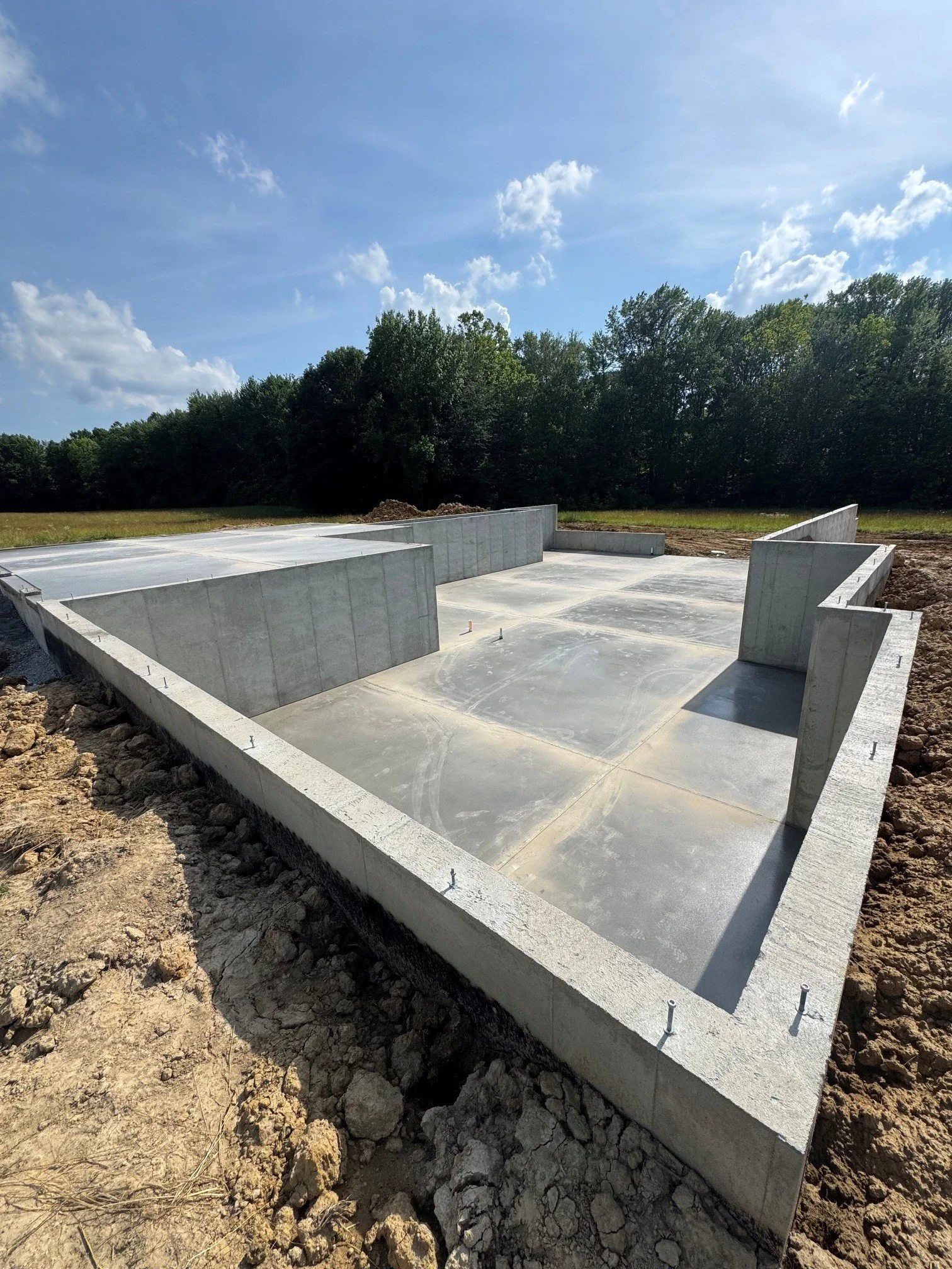 Concrete foundation for a new building under construction on a partly cloudy day, with trees in the background and some dirt and rocks at the site.