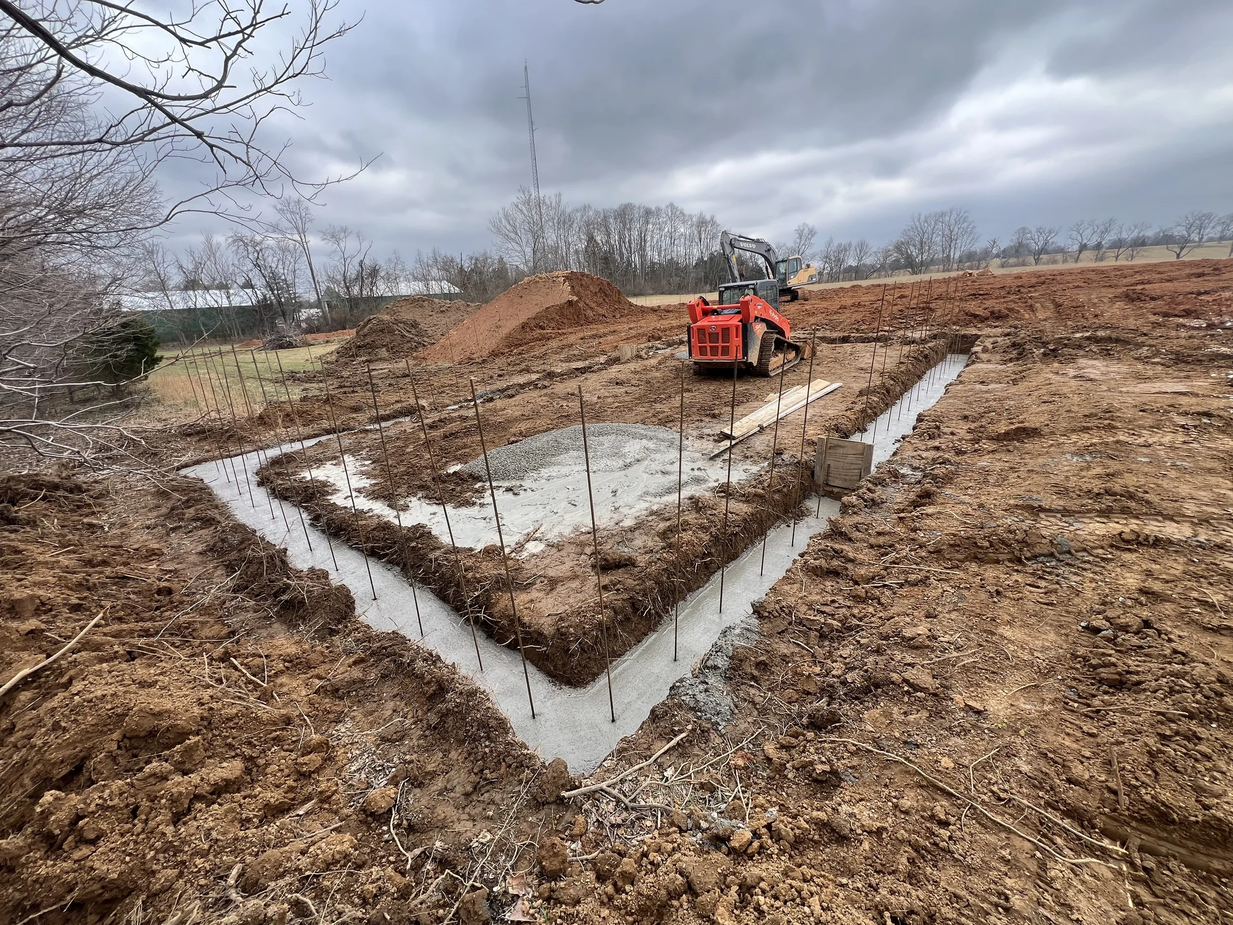 Construction site with excavated trenches, a red compact excavator, and a mound of dirt under a cloudy sky.