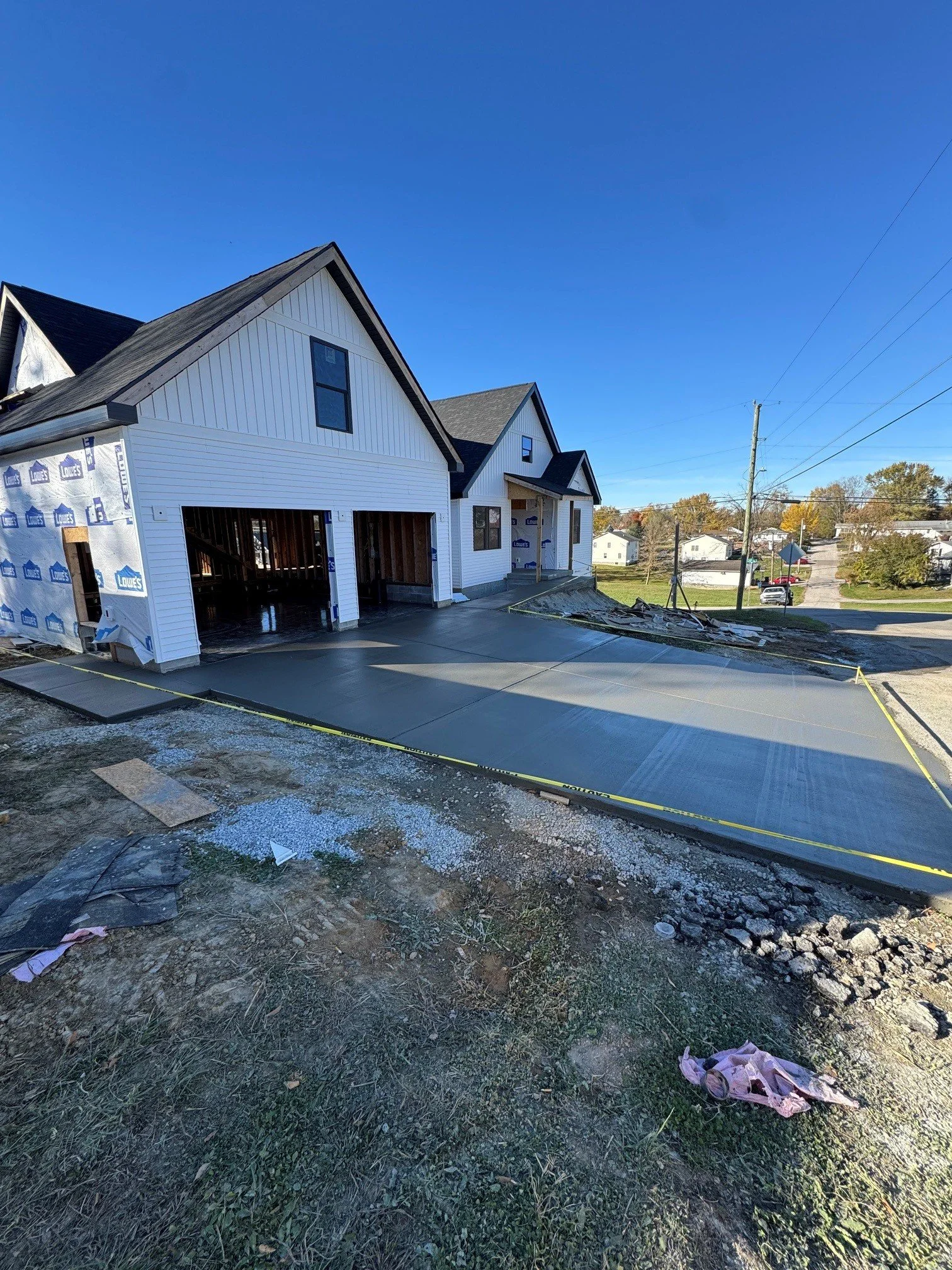 A house under construction with a white exterior and black roof, with two garage openings, set on a newly paved driveway. There is construction debris and dirt in the foreground, and neighborhood houses and trees are visible in the background on a cl