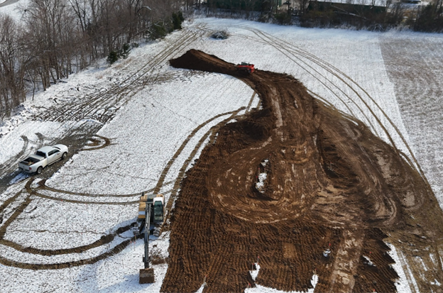 An aerial view of a construction site in a snowy field with tire tracks and a large excavator digging a hole.