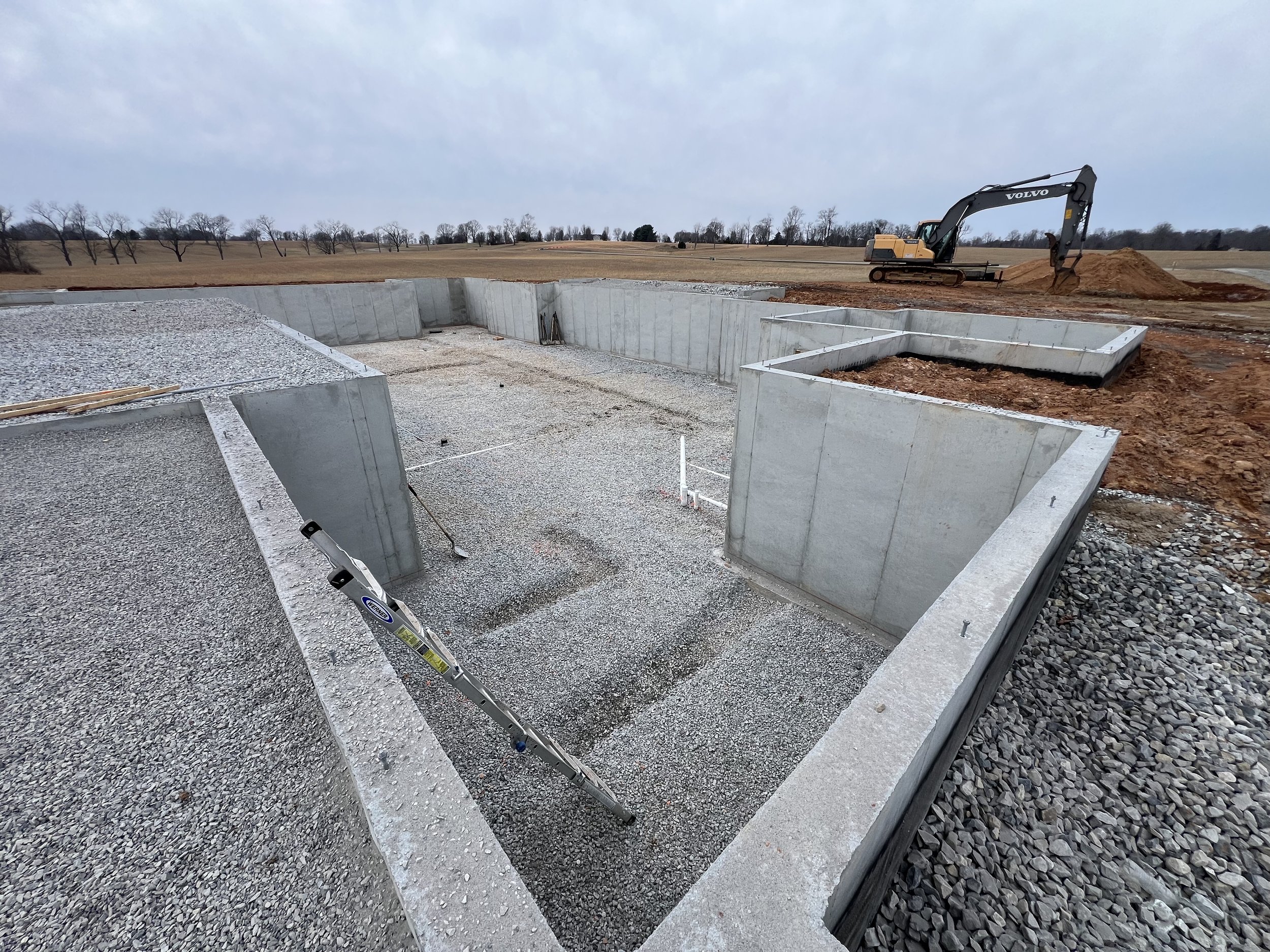 Construction site with concrete foundation, gravel, and a Volvo excavator digging dirt in a rural area with open fields and trees in the background.