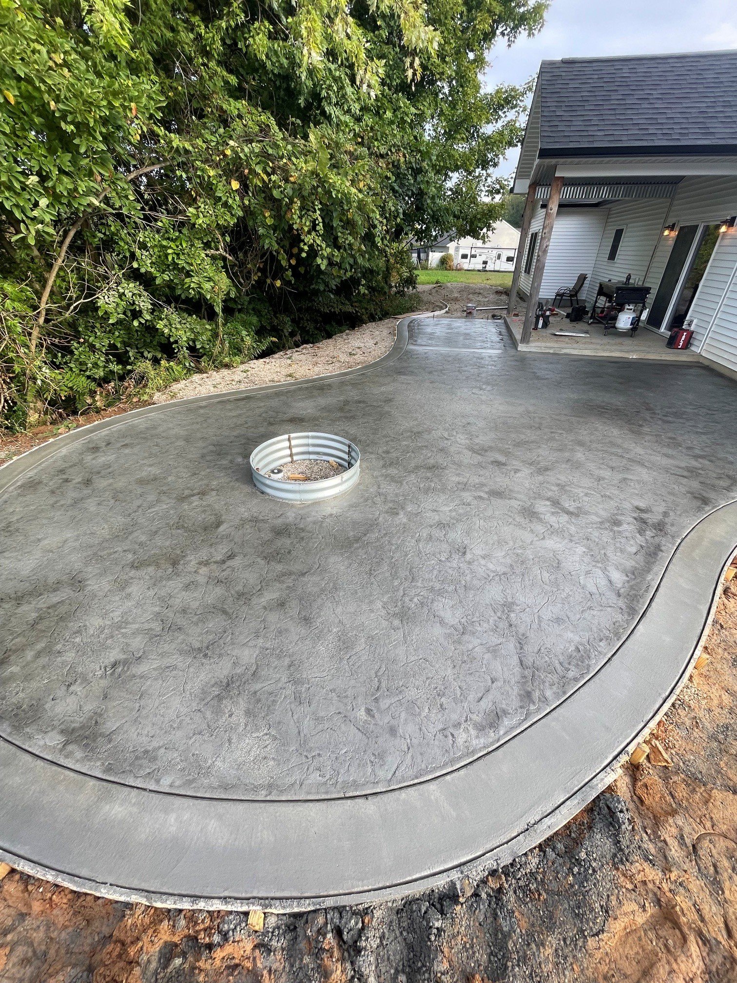 Finished concrete slab in a backyard with a curved edge, next to a house with outdoor furniture and landscaping, surrounded by a tree and a dirt area.
