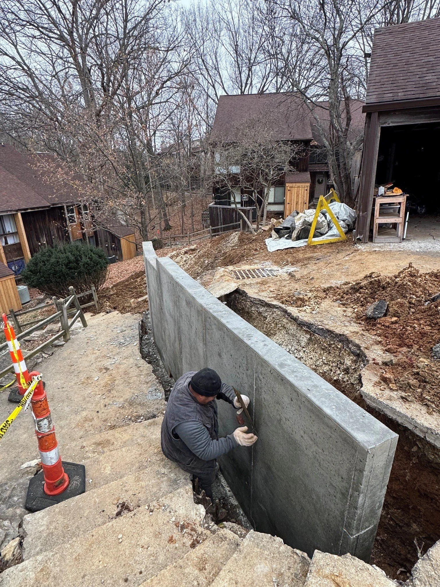 A construction worker installing a large concrete wall outside a residential area, with trees, houses, and construction equipment visible.