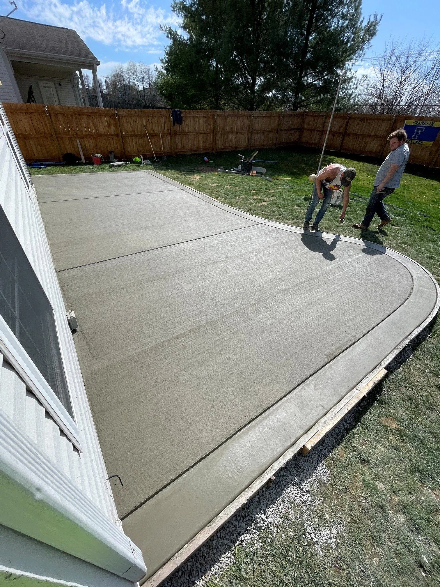 Freshly poured concrete patio with smooth surface and curved edges in a backyard, fenced with new wooden fencing, with two people working on finishing the concrete.