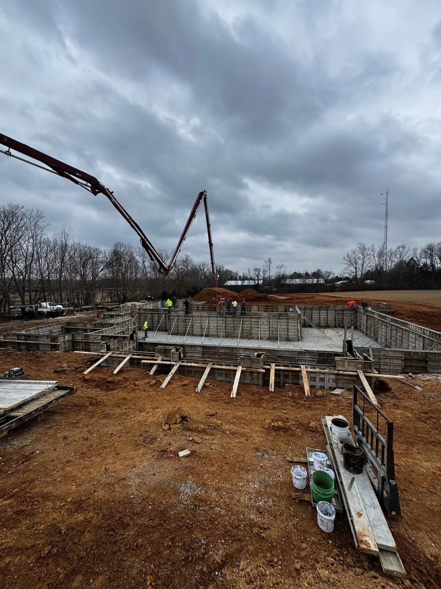Construction site with concrete foundation and workers. Overcast sky with dark clouds. Utility pole in background. Soil and construction materials in foreground.