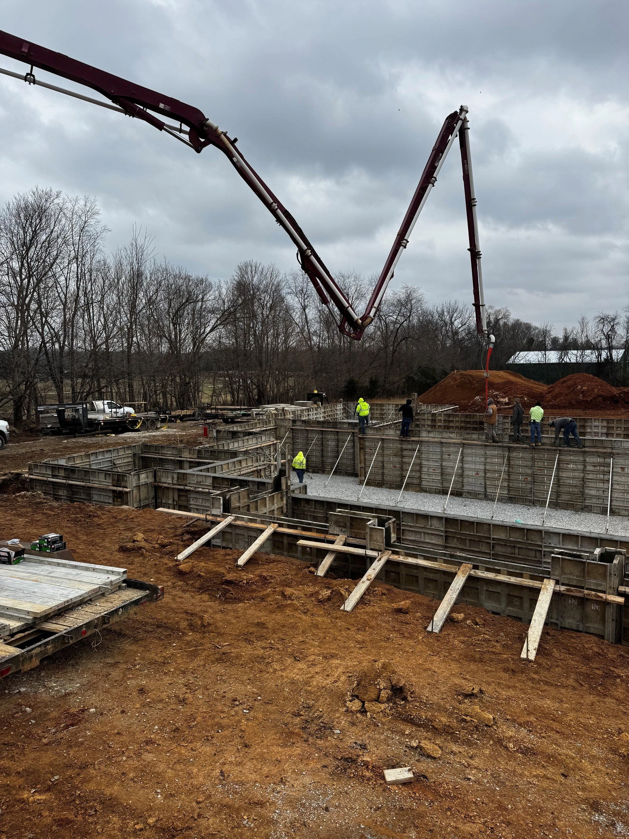 Construction site with concrete footing forms and a concrete pump delivering concrete, workers in safety vests, overcast sky, trees in the background.