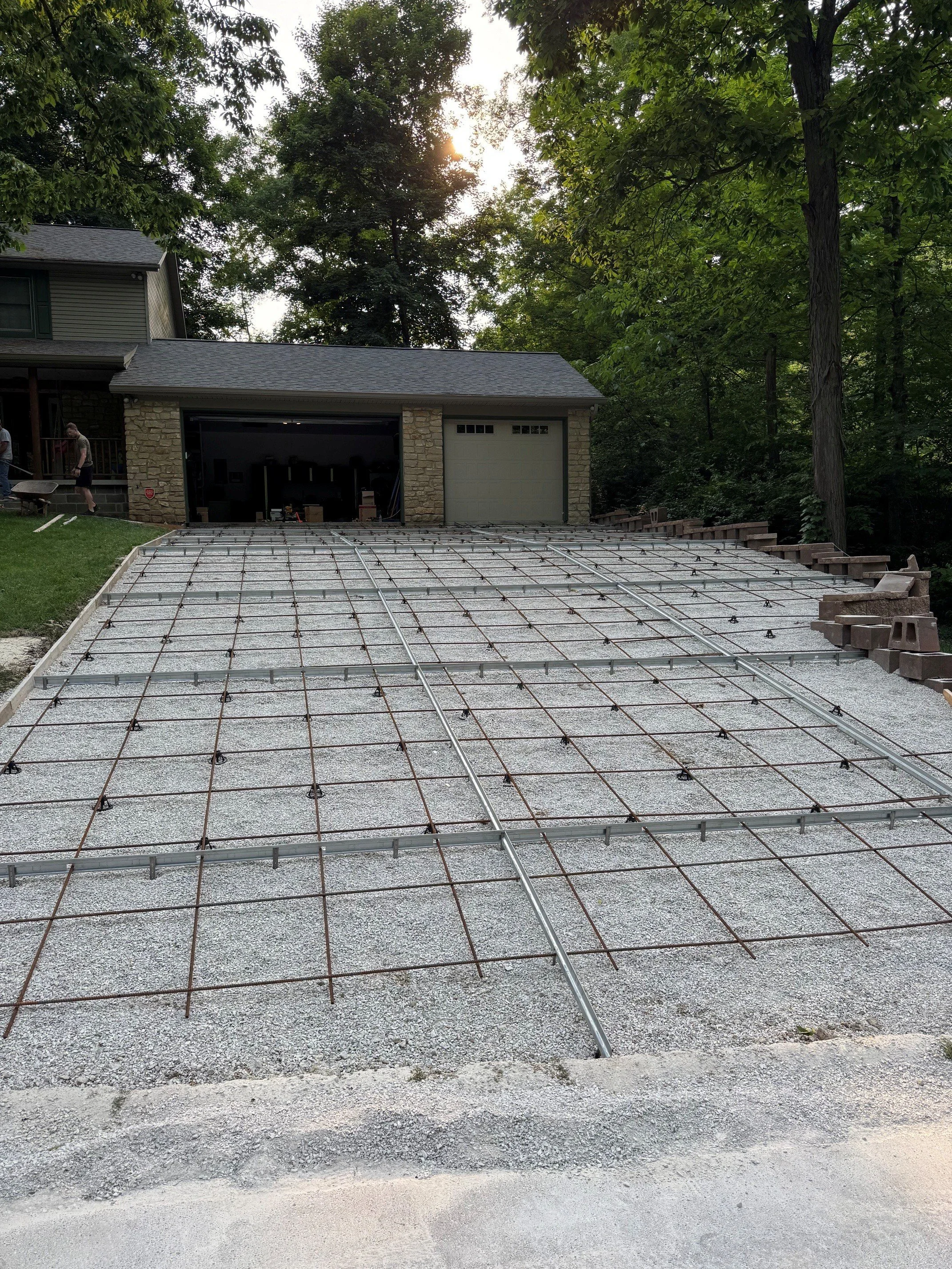 A driveway with a carport or garage in the background, construction materials and rebar in the foreground, workers on the left side, and surrounded by trees and foliage.