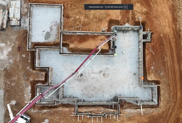 Aerial view of a construction site showing a foundation with a concrete slab and surrounding framing, with a concrete pump truck extending its boom into the area.
