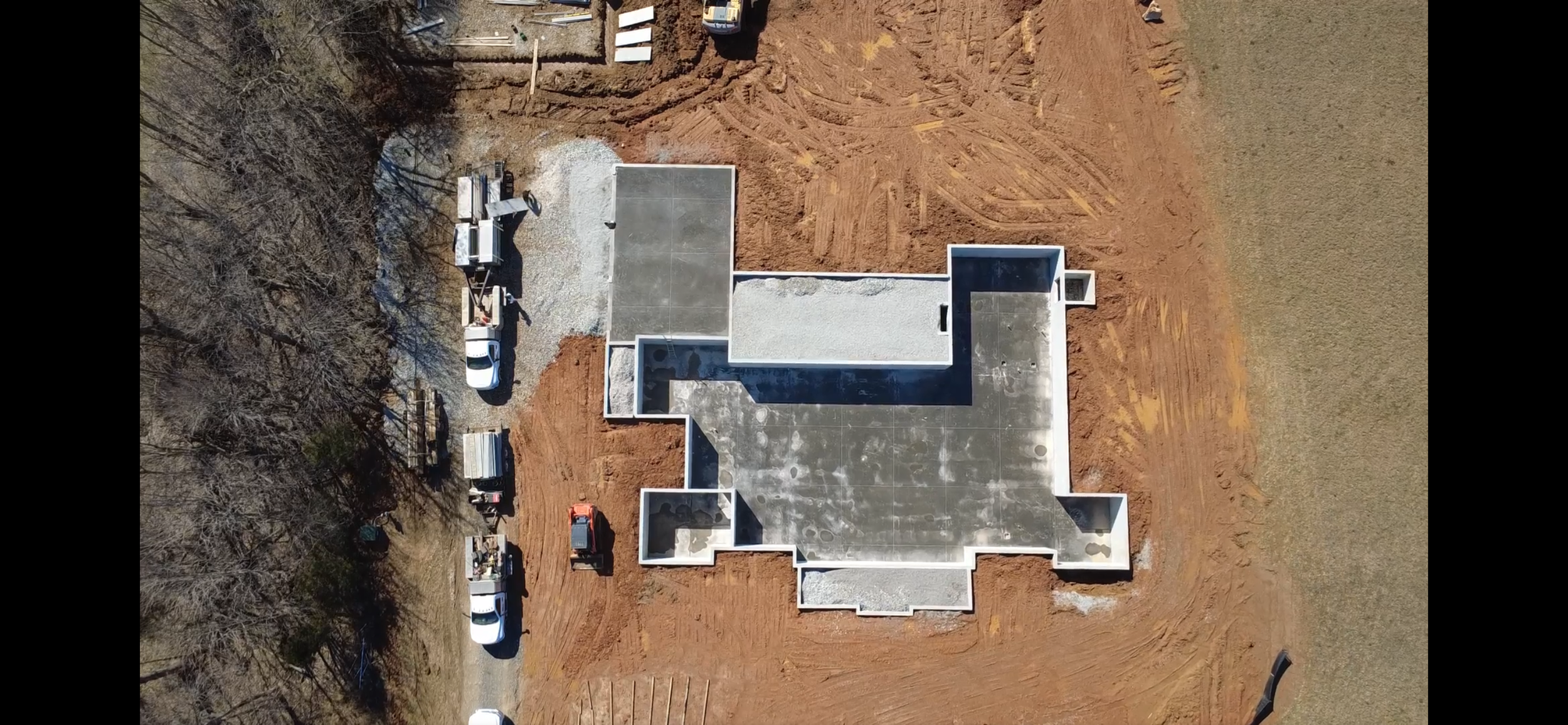 An aerial view of a construction site featuring a building foundation with concrete walls, surrounded by dirt, construction vehicles, and equipment.