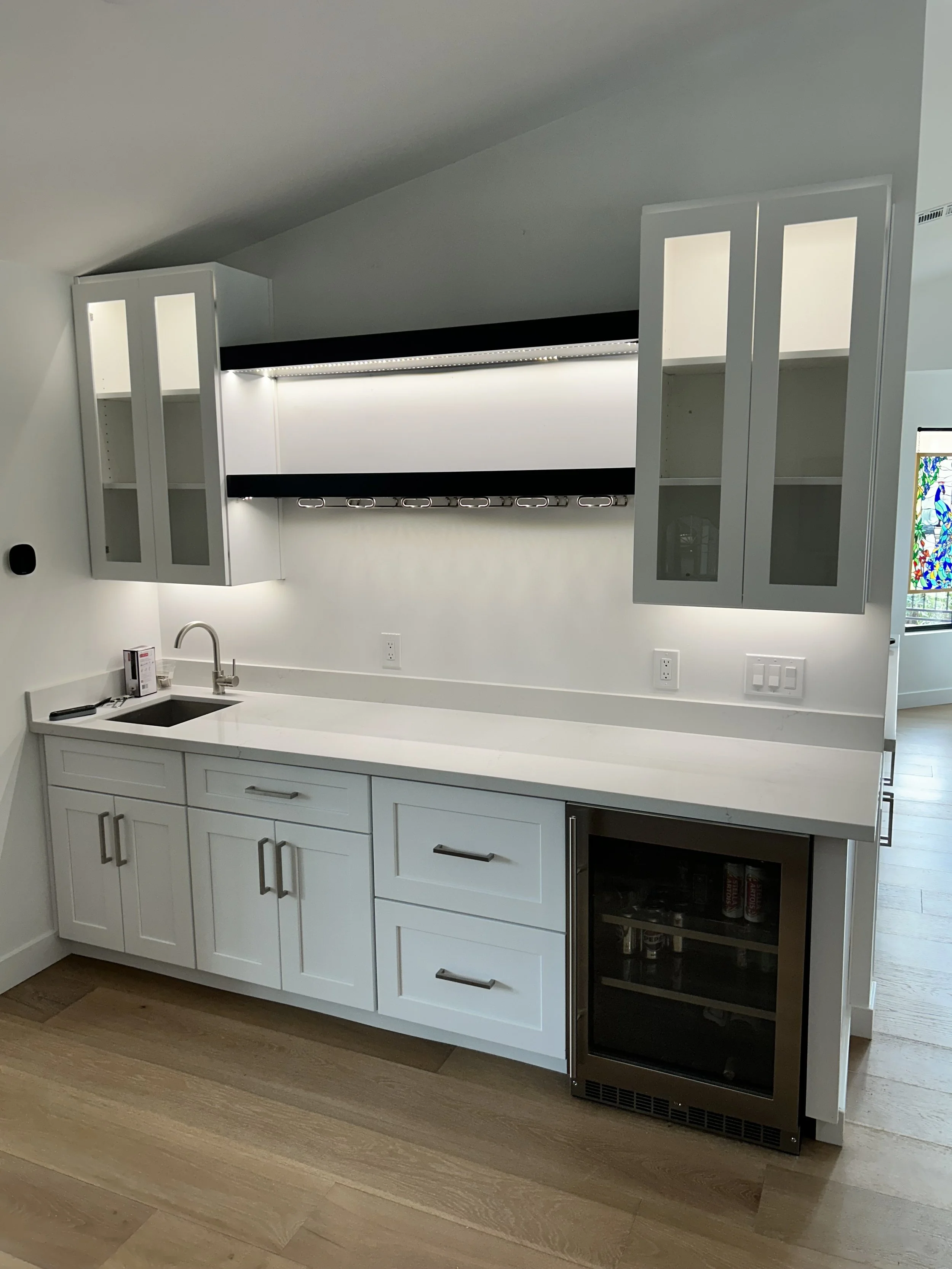 Kitchen with white cabinets, a small sink, a black mini fridge, and wall-mounted cabinets with glass doors and lighting, with hardwood flooring.