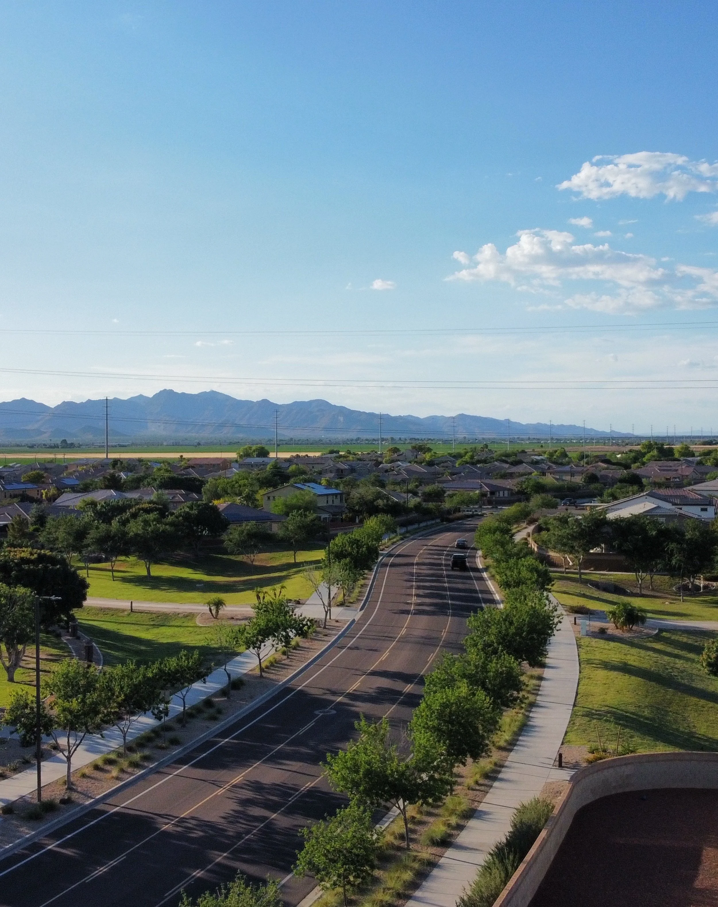 A wide view of a suburban street with a mountain range in the background. Trees line the sidewalks, and houses are visible along the street. The sky is blue with some clouds.