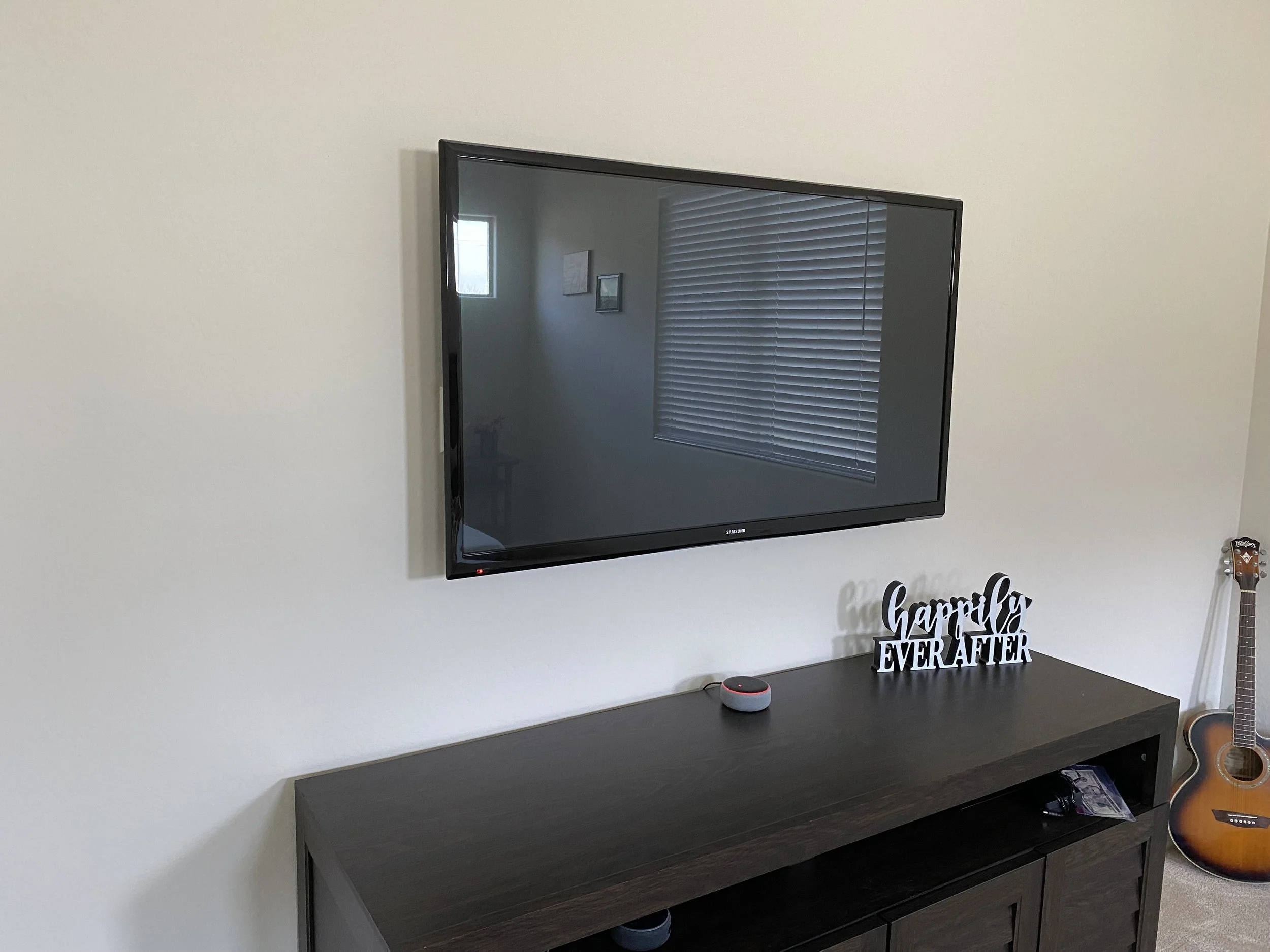 Living room with wall-mounted TV, window with blinds, decorative wall art, a countertop with a smart speaker, a sign that says "happily ever after", and two acoustic guitars, one brown and one tan.