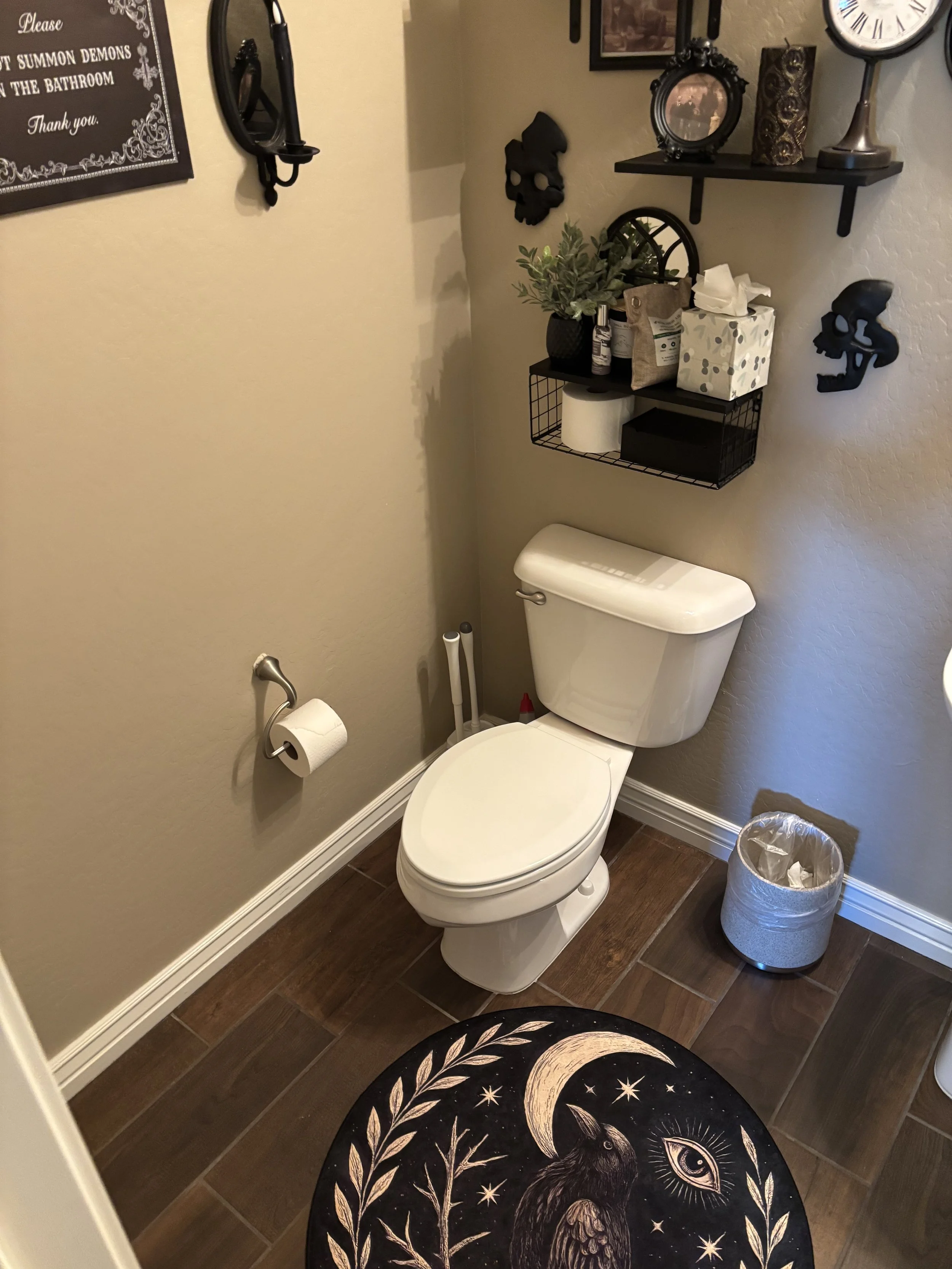 A small bathroom with beige walls and dark wood flooring. There is a white toilet with the lid open, and a round rug featuring a black moon and stars design in front of it. Above the toilet is a black shelf holding toiletries, tissues, and decorative