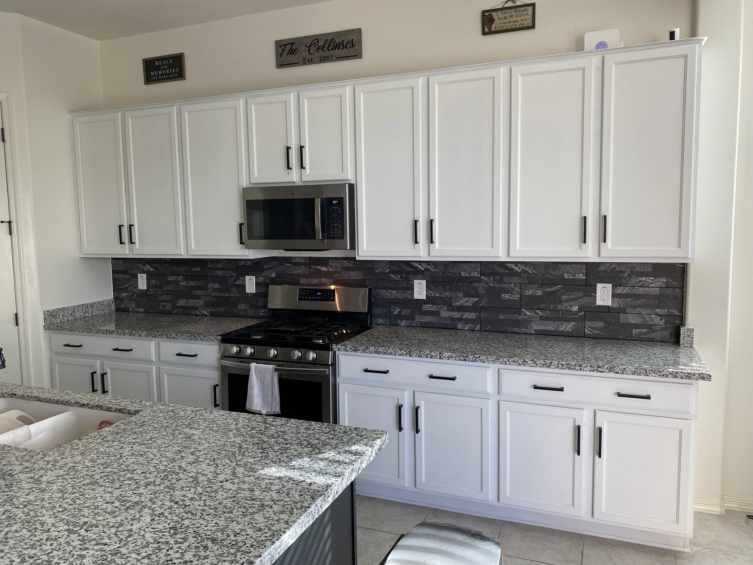 Modern kitchen with white cabinets, granite countertops, black handles, stainless steel microwave, and stove. Decorative wall signs above cabinets, including a wooden sign that reads 'The Collinces Est. 2007'. The backsplash is made of black textured