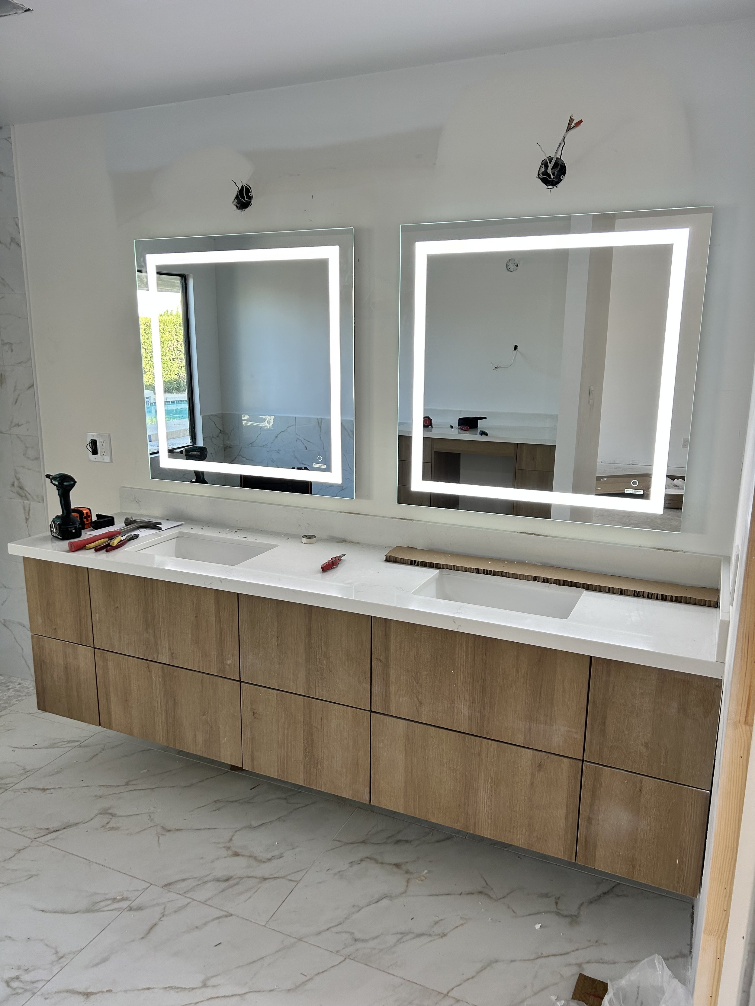 Bathroom vanity with two illuminated mirrors, wooden cabinets, white marble countertop, tools, and partially installed electrical wiring.
