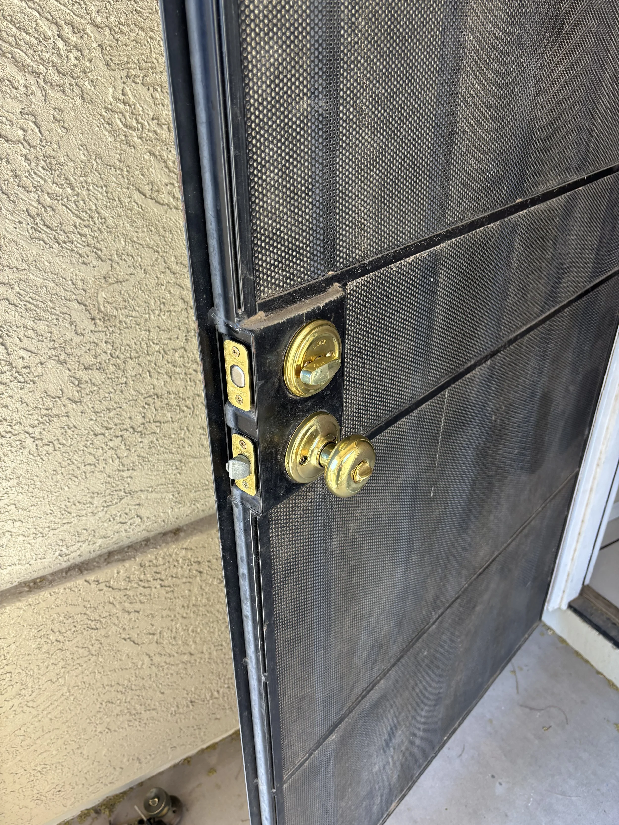 Close-up of a black metal screen door with a gold deadbolt lock and a doorknob in front of a beige textured wall.