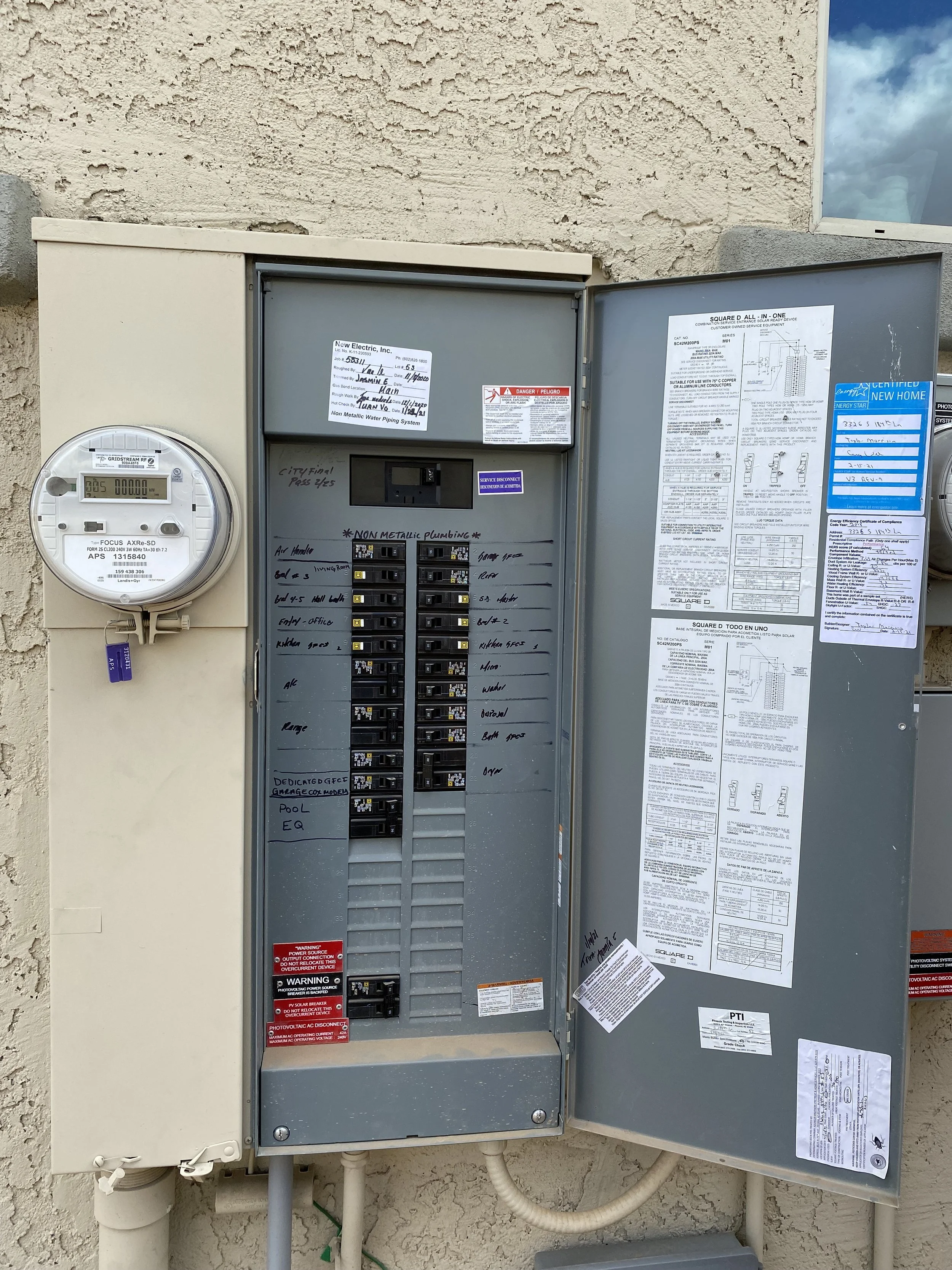 An electrical panel with various labels and documents attached, mounted on an exterior wall.