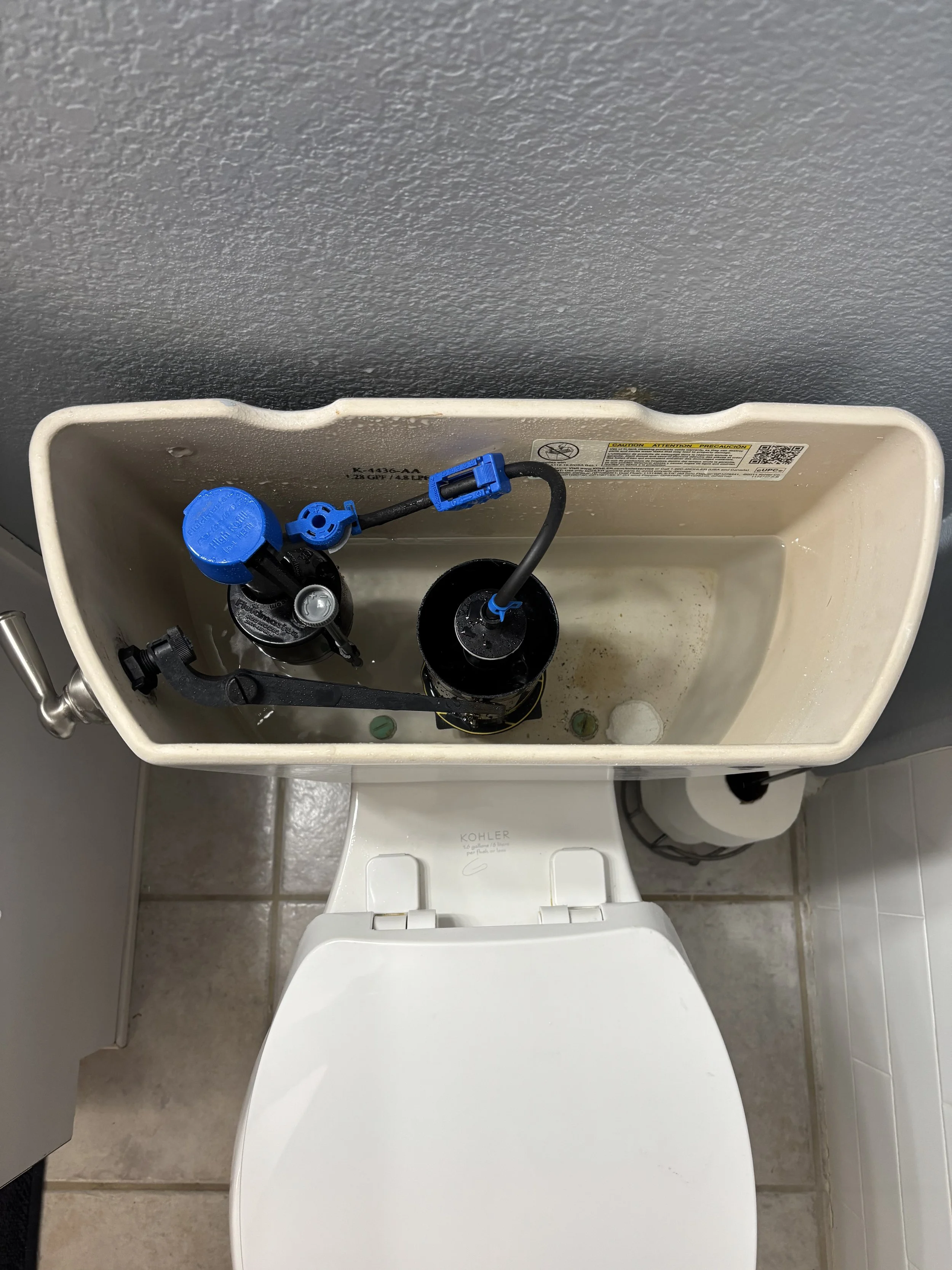 The inside of a toilet tank with its lid removed, showing various components including a blue fill valve, a flush valve, and fluid inside the tank. The tank is white and mounted on a white toilet base in a bathroom with tiled floor.