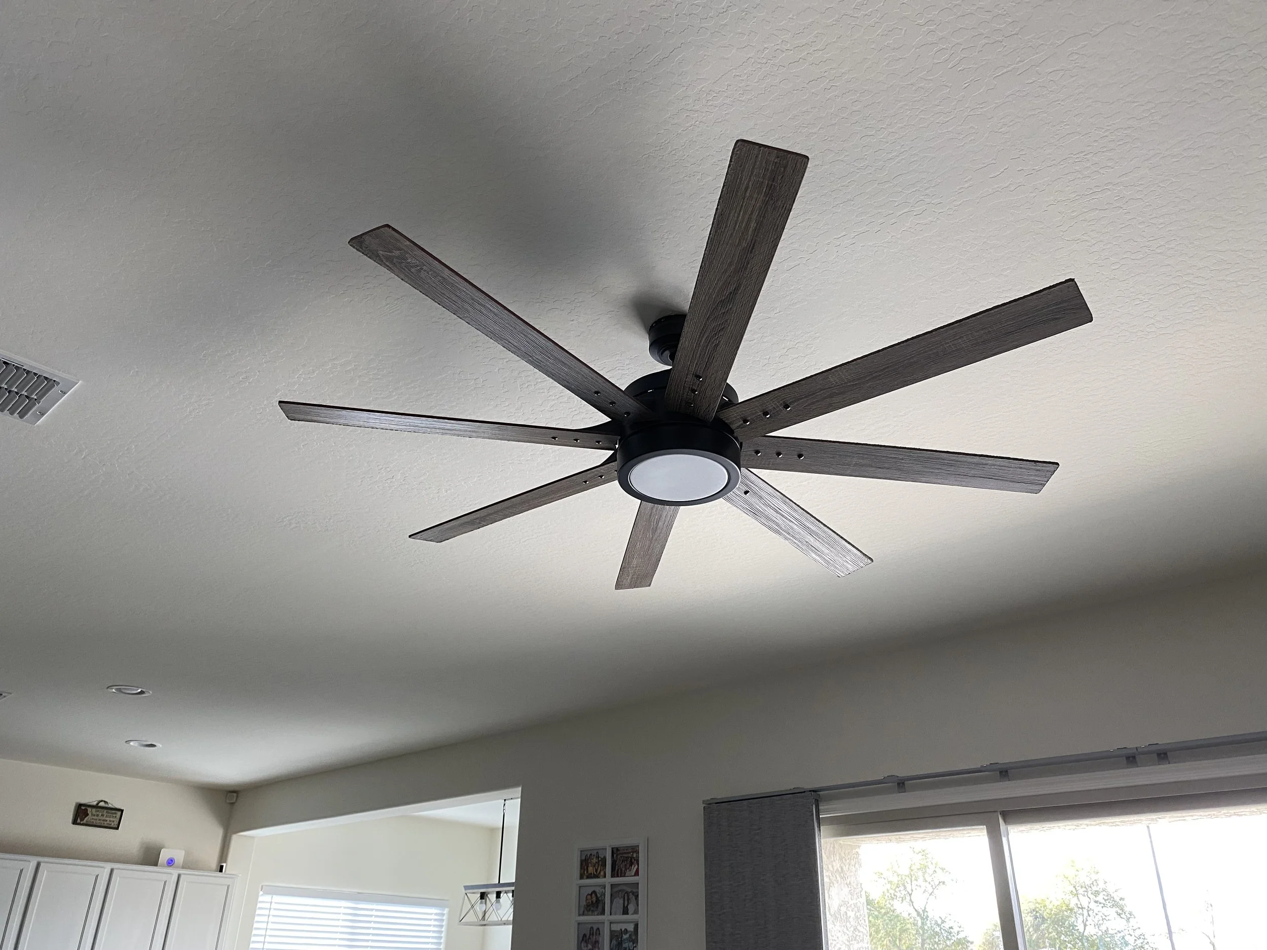 Ceiling fan with wooden blades and a light fixture in a living room with windows and white cabinets.