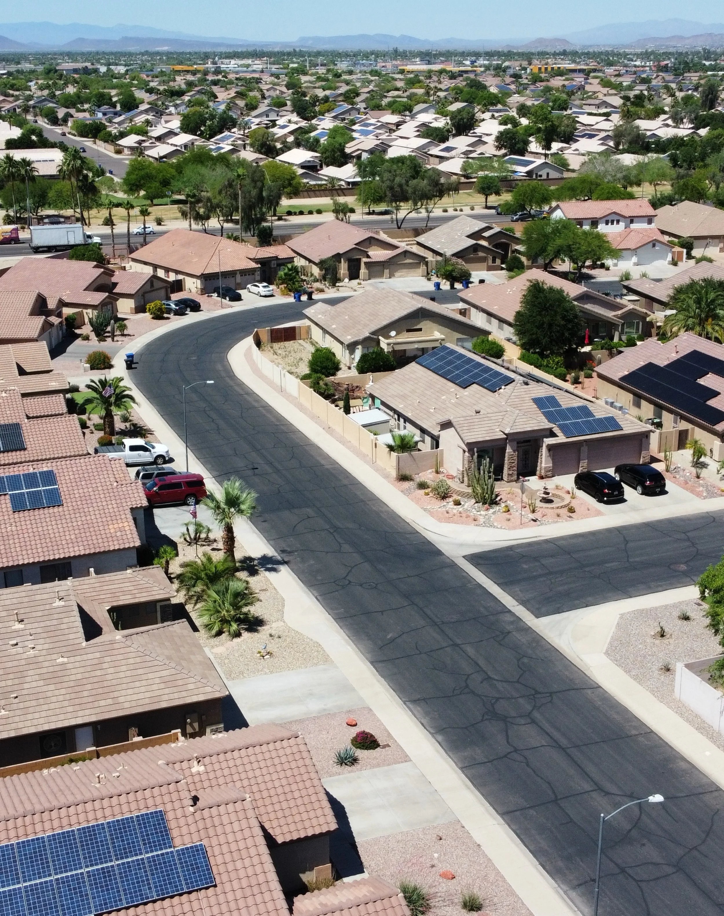 An aerial view of a suburban neighborhood featuring single-story houses, many with solar panels on the roofs, surrounded by trees, with a curving street, parked cars, and desert landscaping. In the background, mountains are visible under a clear sky.
