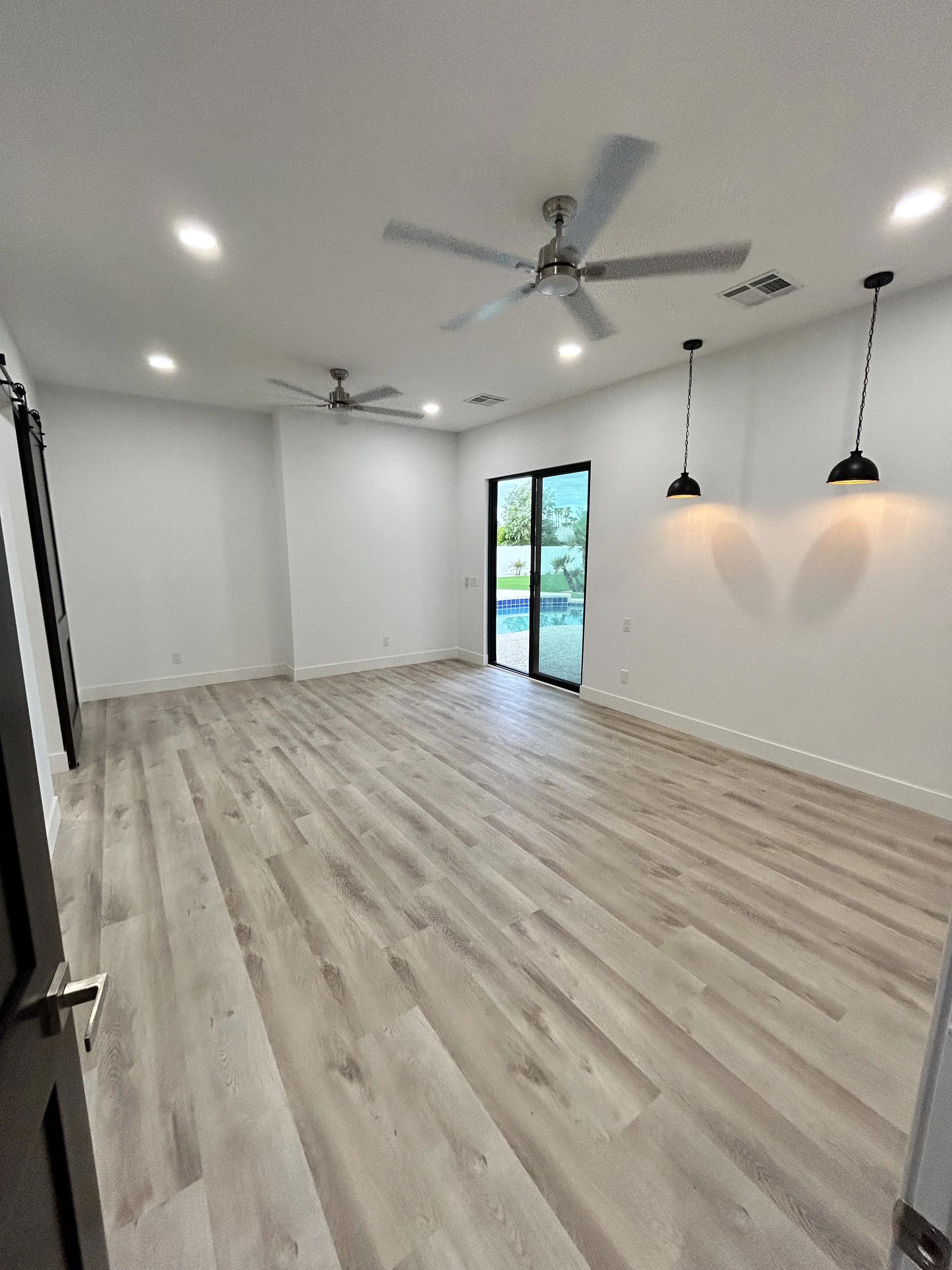 Empty living room with light wood floors, white walls, ceiling fans, black pendant lights, sliding glass door showing outdoor pool, trees, and sky.