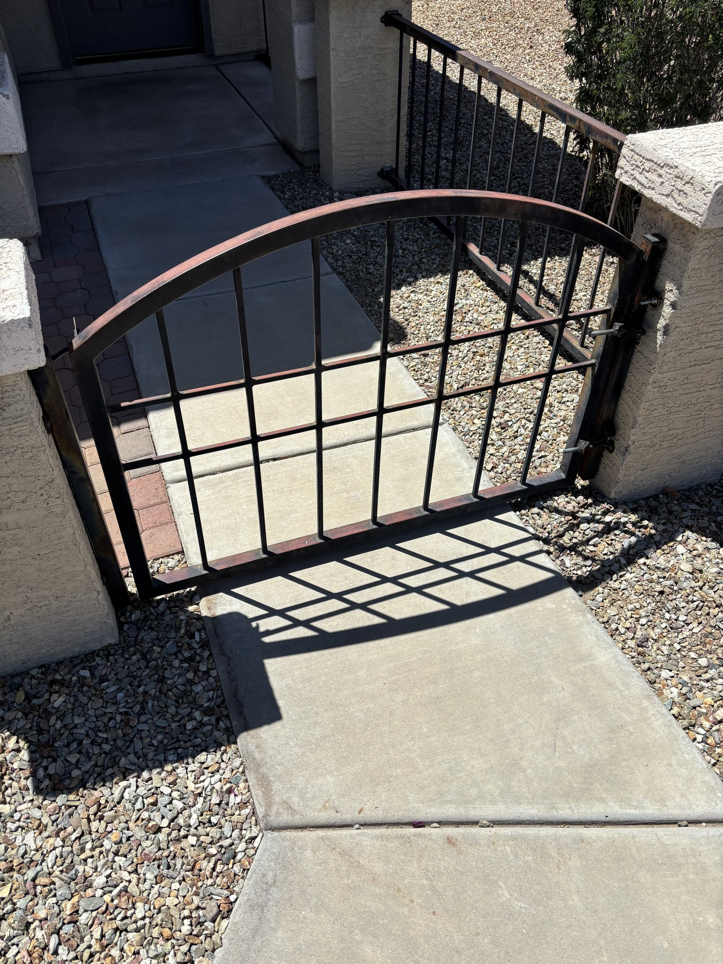 A small metal gate with a curved top, attached to a brick column next to a walkway, casting a shadow on the concrete sidewalk.
