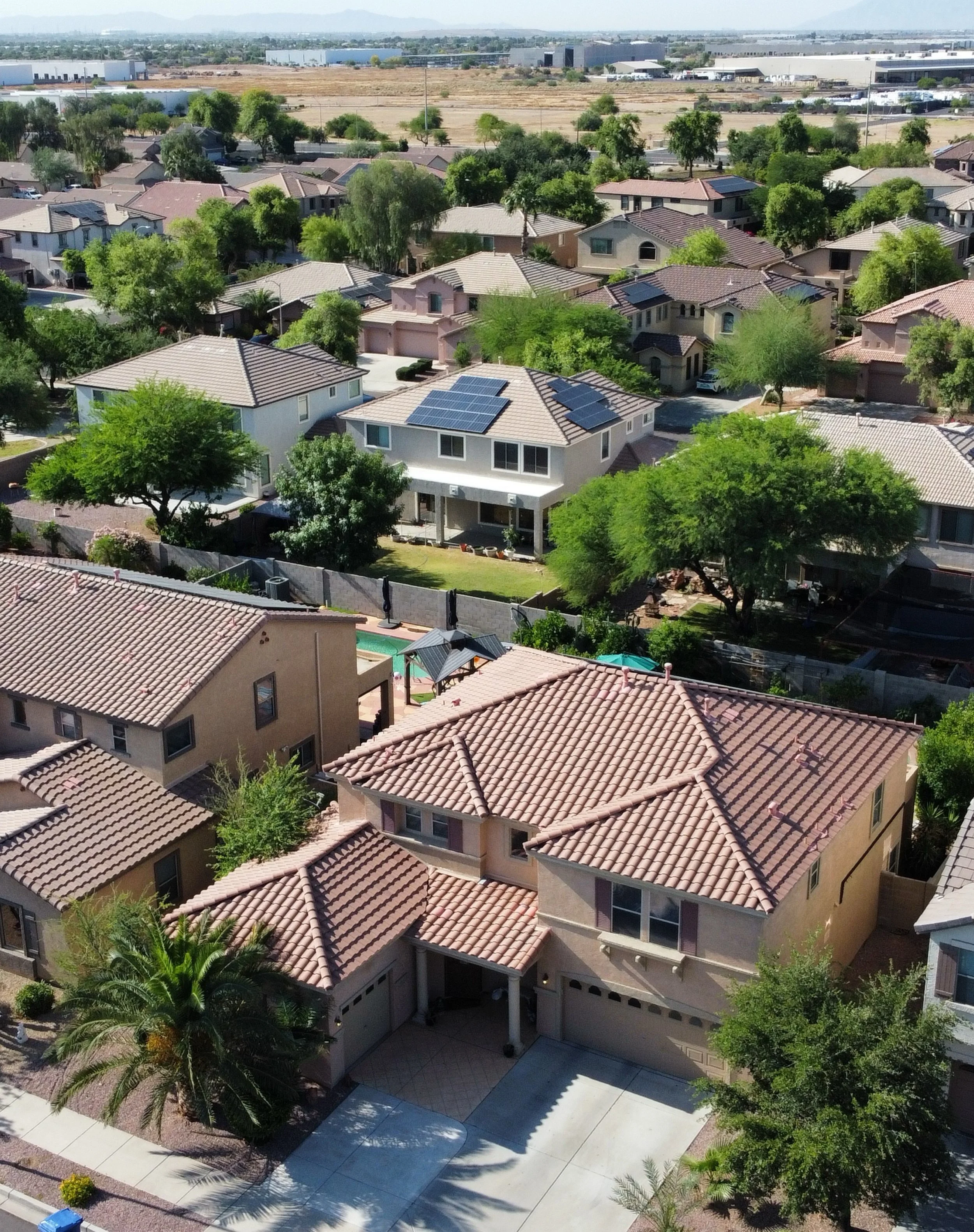 Aerial view of a residential neighborhood with multiple houses featuring tile roofs, some with solar panels, surrounded by trees, with a commercial or industrial area in the background.