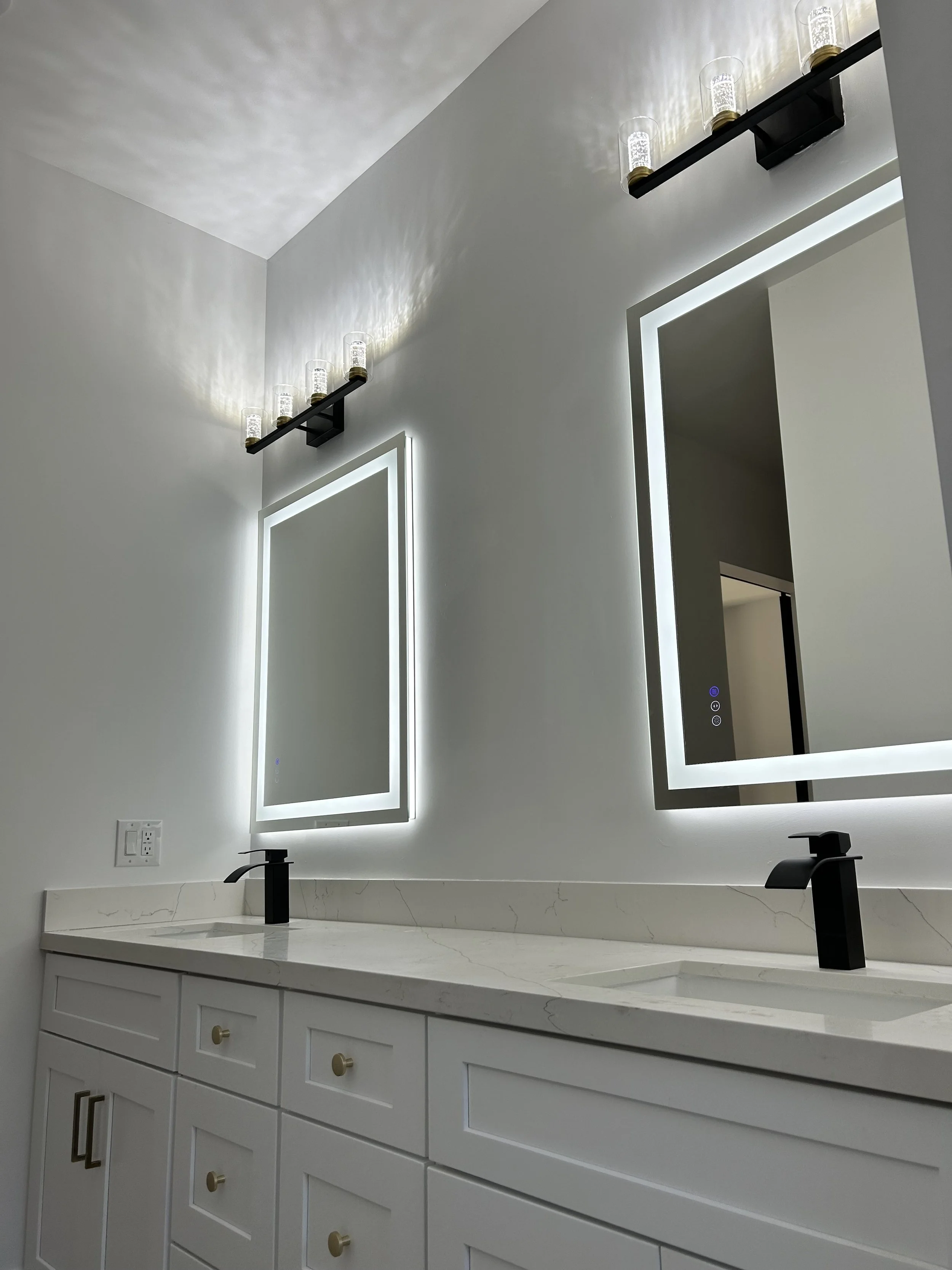 Modern bathroom vanity with white marble countertop, two black undermount sinks with black faucets, illuminated mirror, and a black shelf with five glass candle holders with candles.