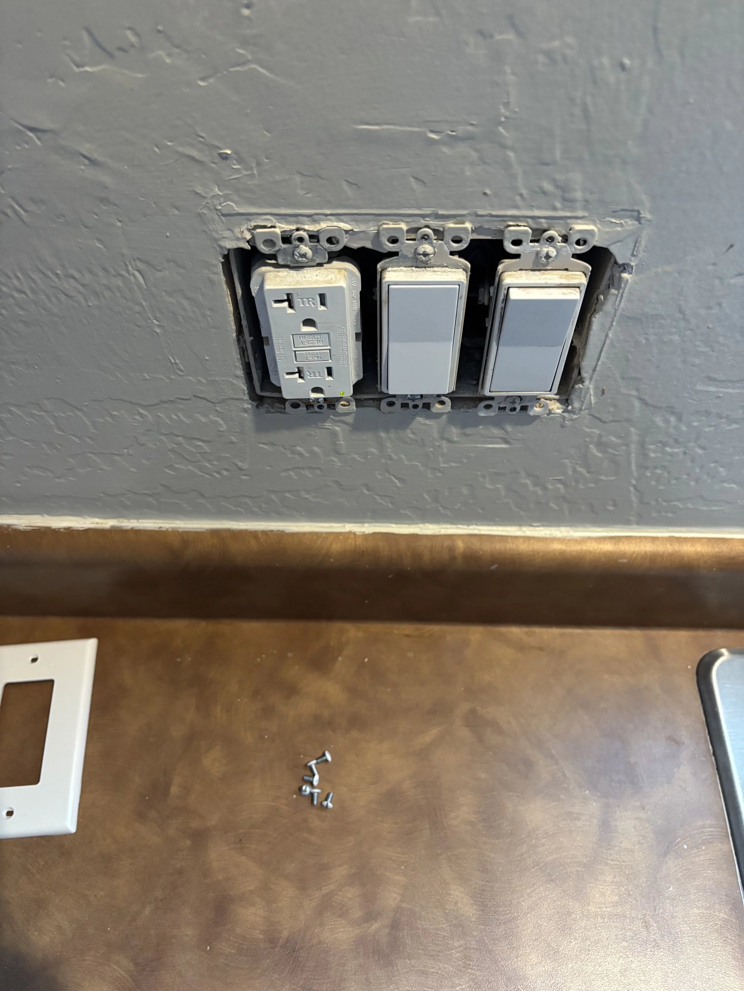 An electrical outlet, two white cover plates, and scattered screws and nuts on a brown wooden surface.