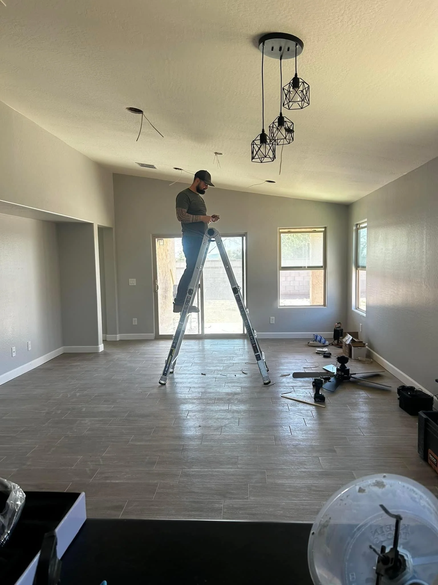 A man standing on a ladder working on a ceiling light fixture in a living room with large windows, hardwood floors, and a gray wall.