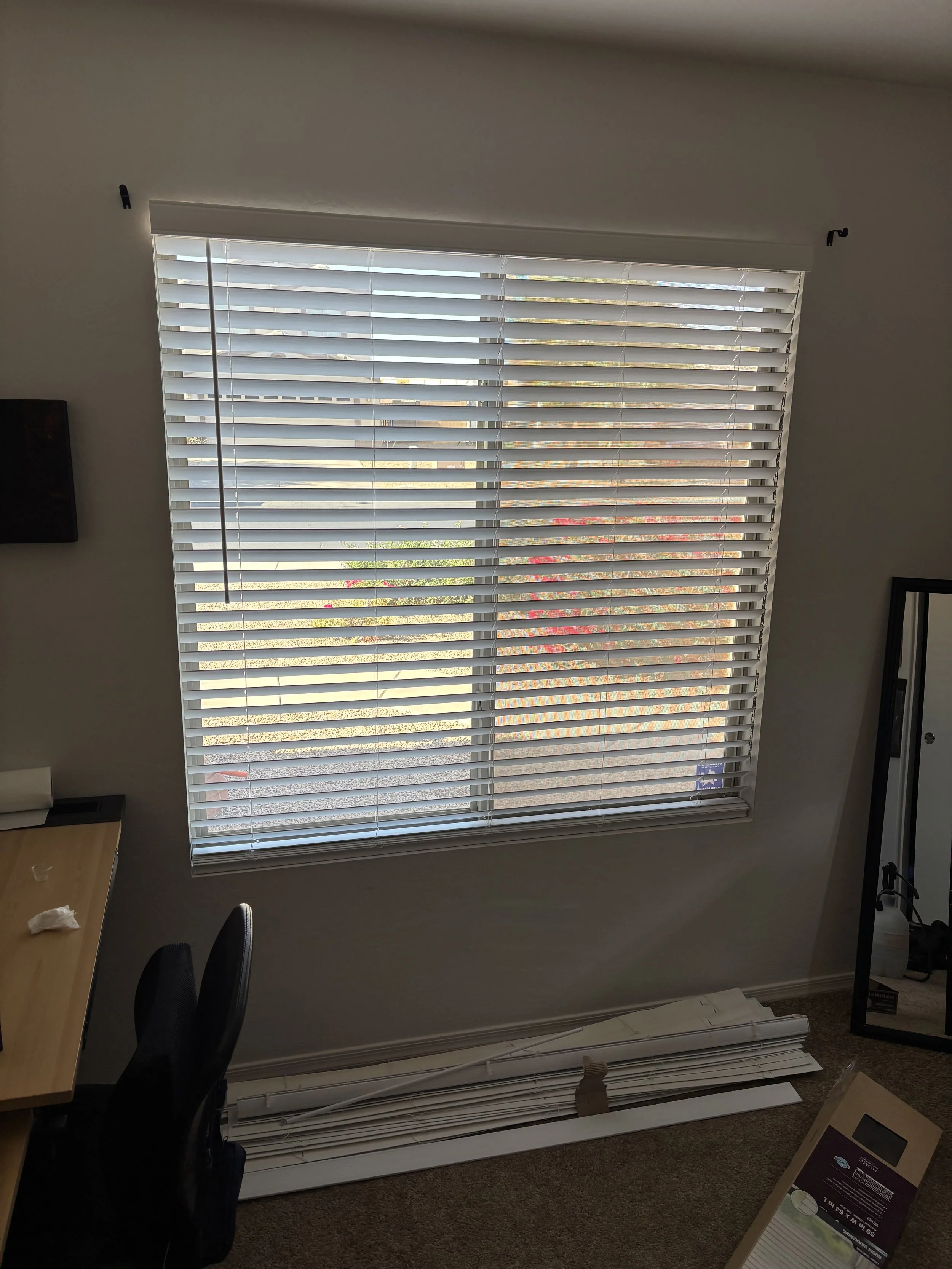 A room with a window covered by white horizontal blinds, with some blinds laying on the floor beneath the window, and a cardboard box on the carpeted floor nearby.