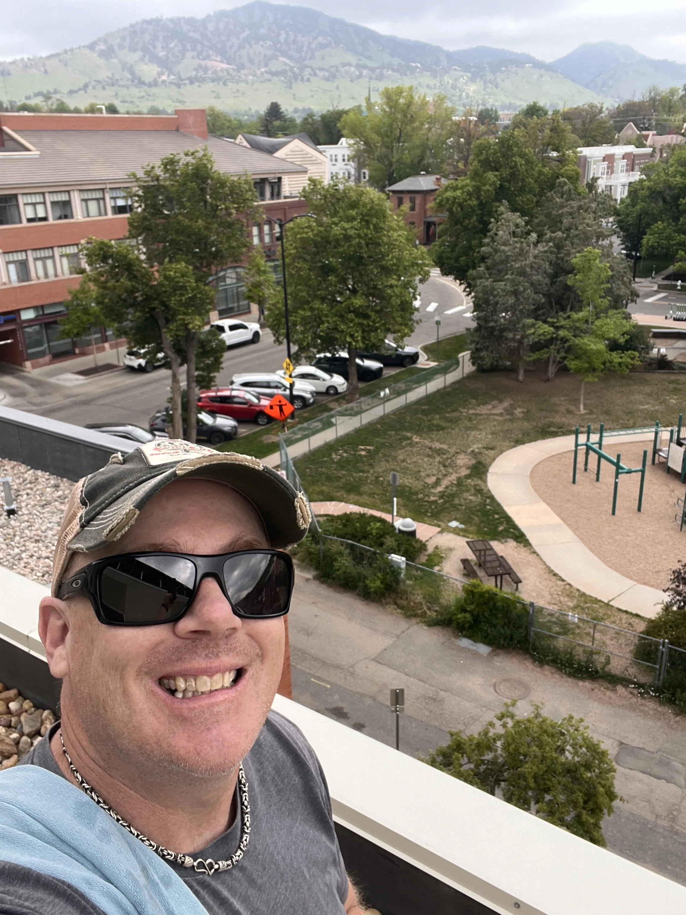A person smiling at the camera wearing sunglasses, a baseball cap, a chain necklace, and a gray t-shirt, outdoors on a balcony with a view of trees, parked cars, and buildings in the background, with mountains in the distance.