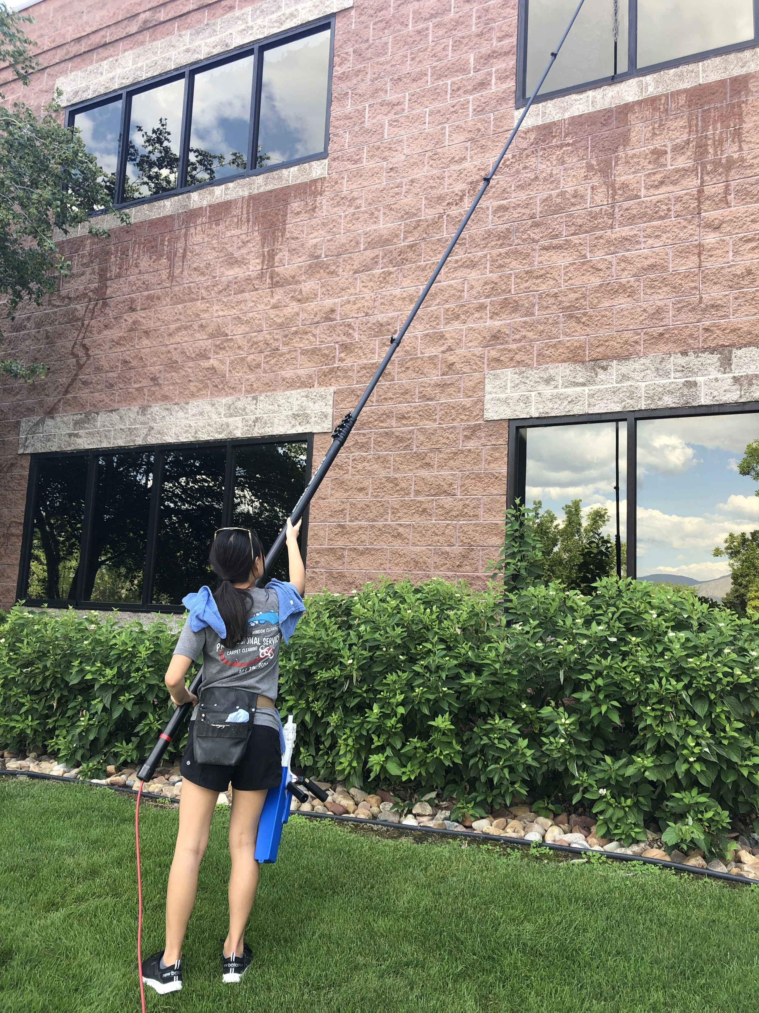 A woman vacuuming outside of a brick building with large windows, green bushes, and a grassy lawn.