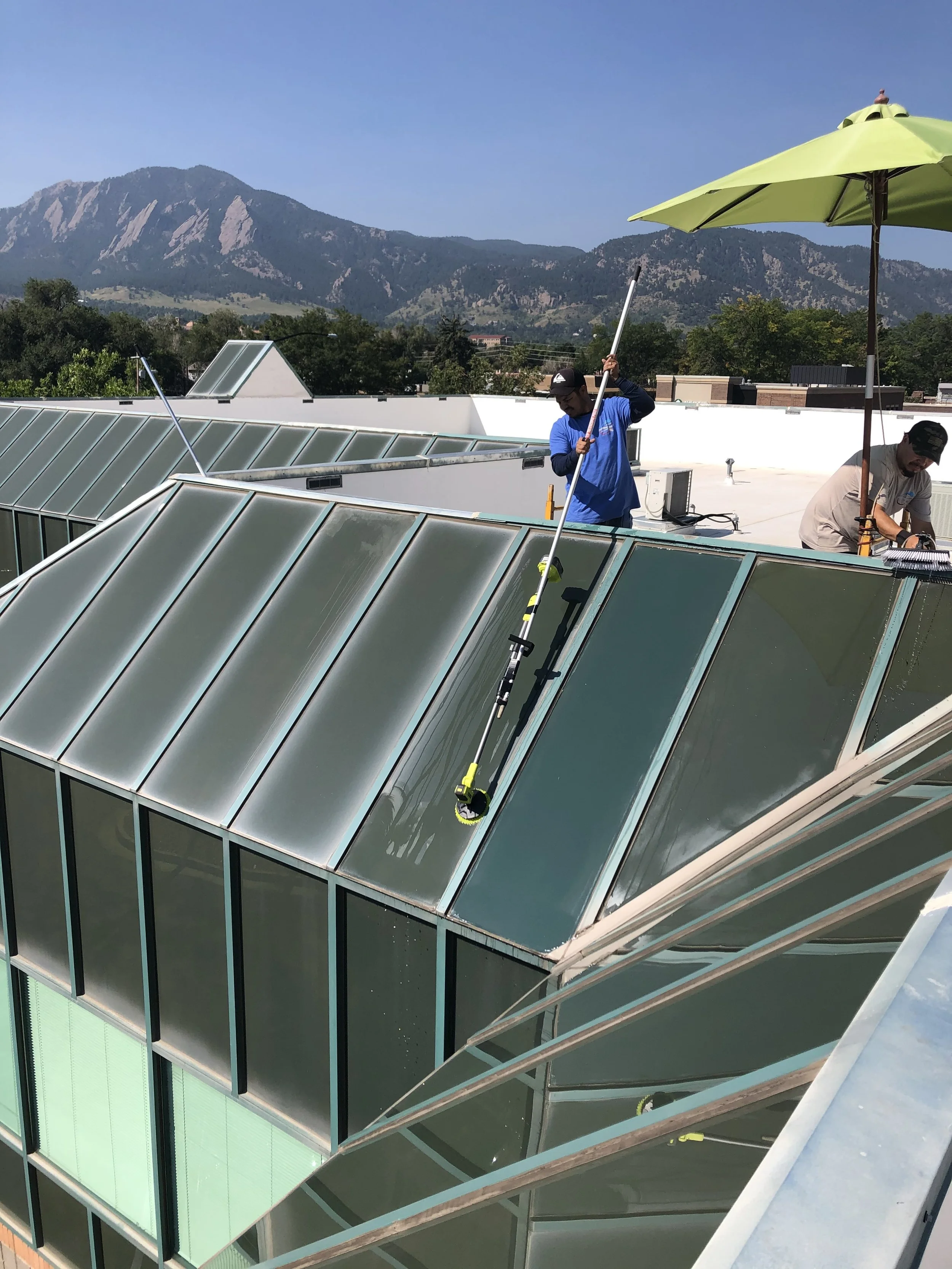 Two workers cleaning the windows of a modern building with glass walls and a sloped roof, with mountains in the background.