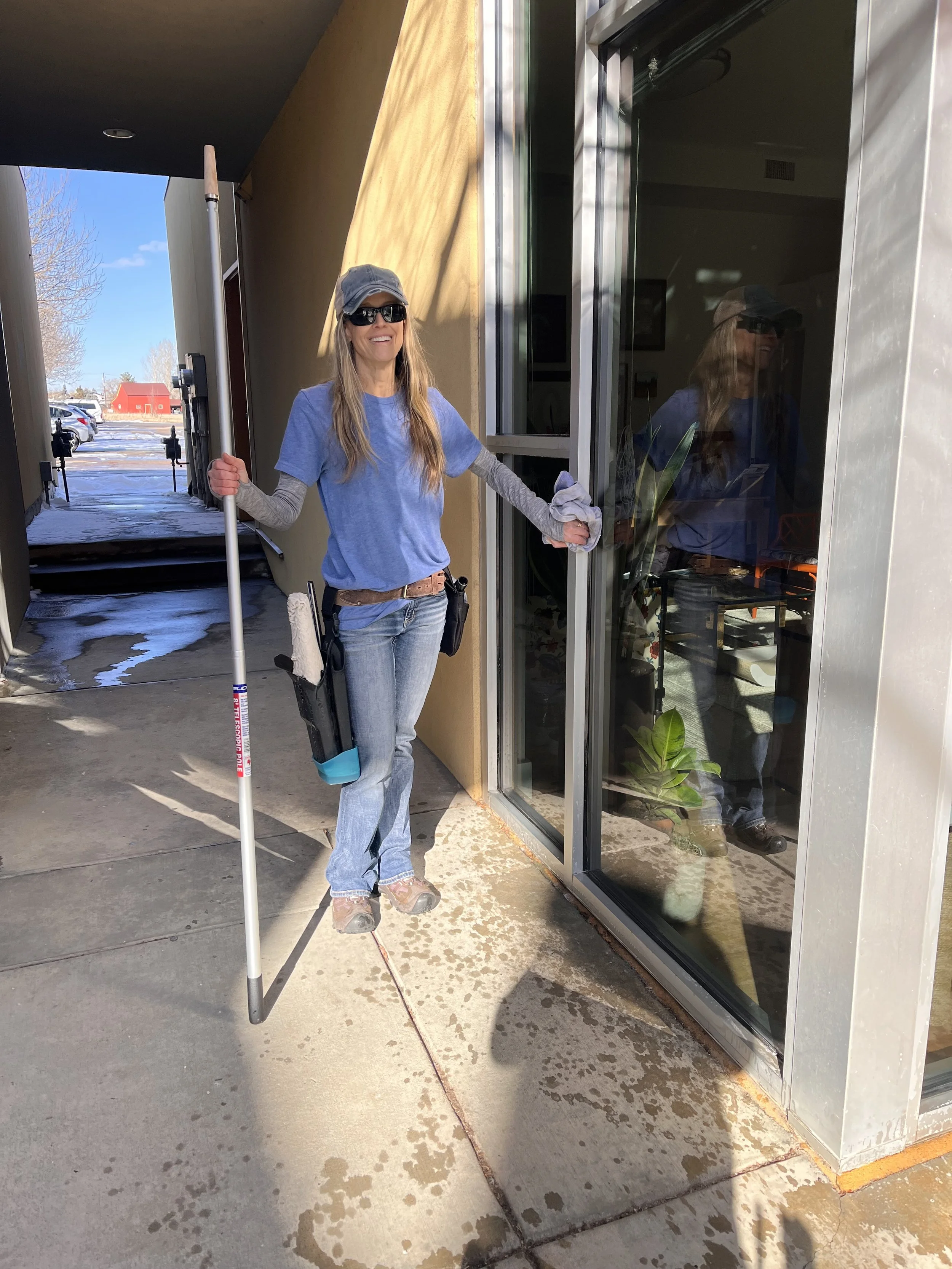 A woman outside a building, smiling, wearing a baseball cap, sunglasses, a gray long-sleeve shirt under a blue T-shirt, jeans, and roller skates, holding a white stick, with a black pouch hanging from her waist, standing near a glass door.
